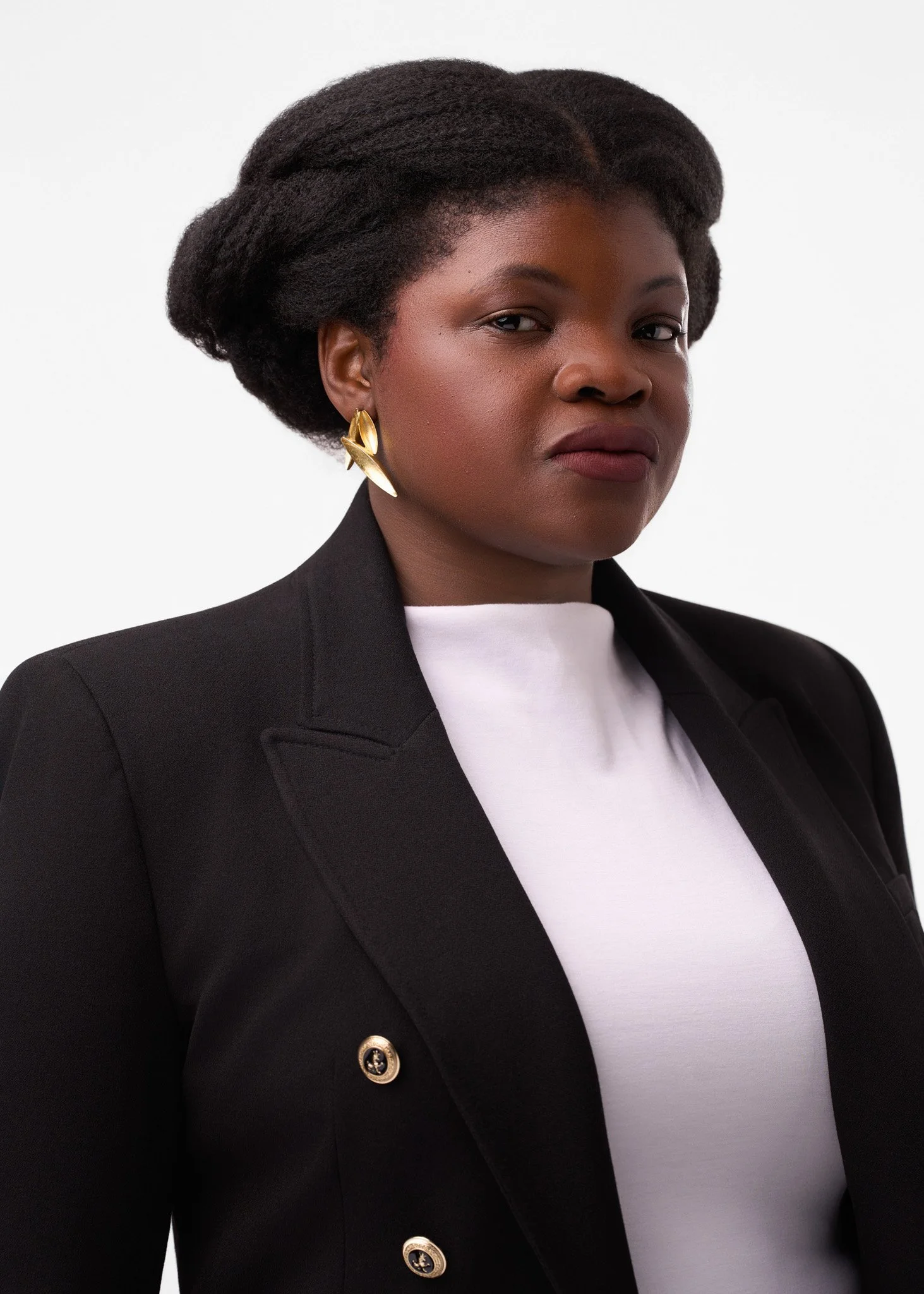 A woman with dark skin, wearing a black blazer with gold buttons, a white top, and large gold earrings. She has short, natural hair styled in a voluminous updo, posing against a plain white background.