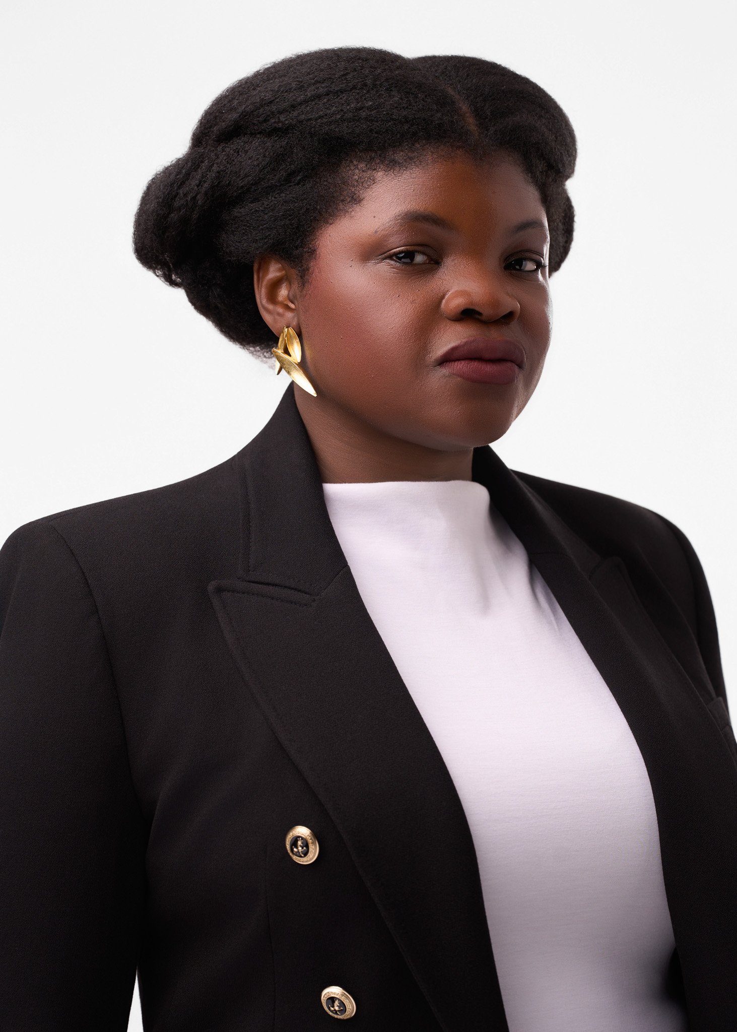 A woman with dark skin, wearing a black blazer with gold buttons, a white top, and large gold earrings. She has short, natural hair styled in a voluminous updo, posing against a plain white background.