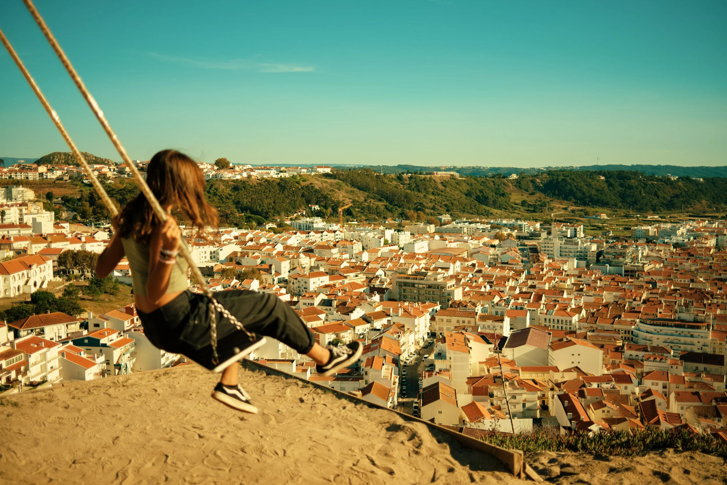 Travel lifestyle image of a person on a swing overlooking a hillside city, wide scenic view with warm light and expansive urban landscape.