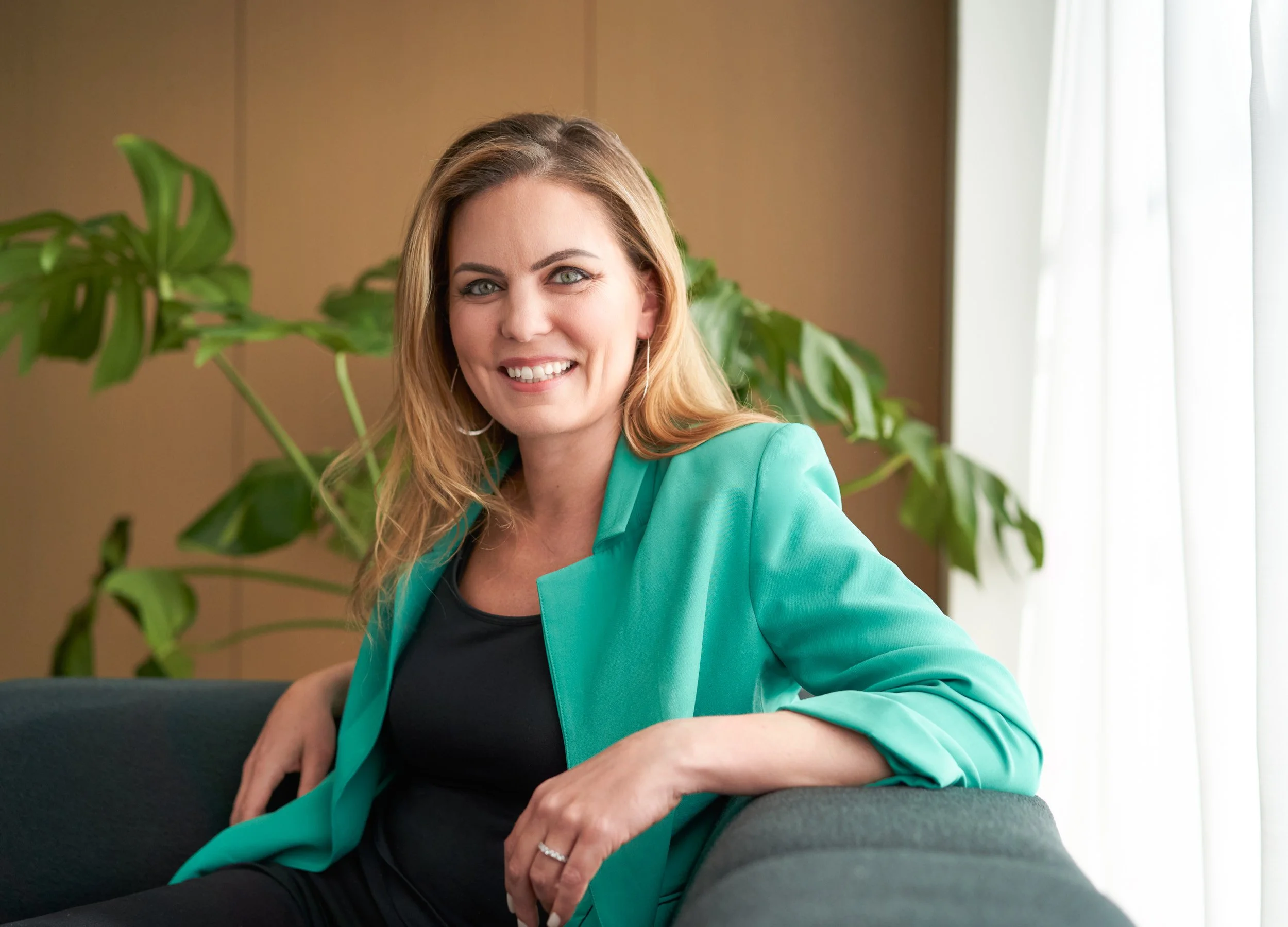 Smiling woman in a green blazer seated in a warm office lounge with a monstera plant, corporate lifestyle portrait photographed in New York City
