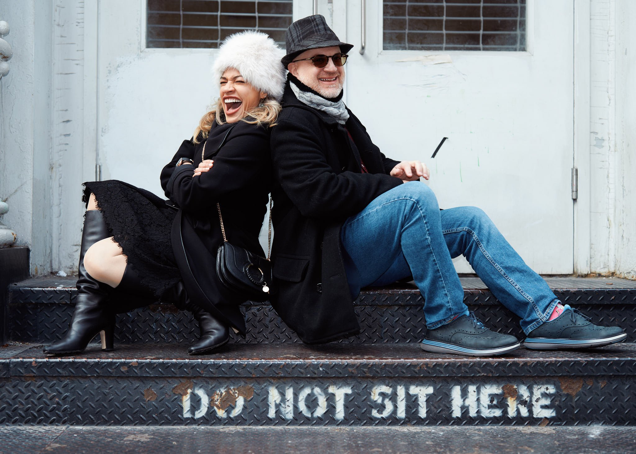 Environmental portrait of two people seated on a metal staircase in New York City, photographed in a relaxed, candid moment with 'DO NOT SIT HERE' painted on it, in front of a white door.