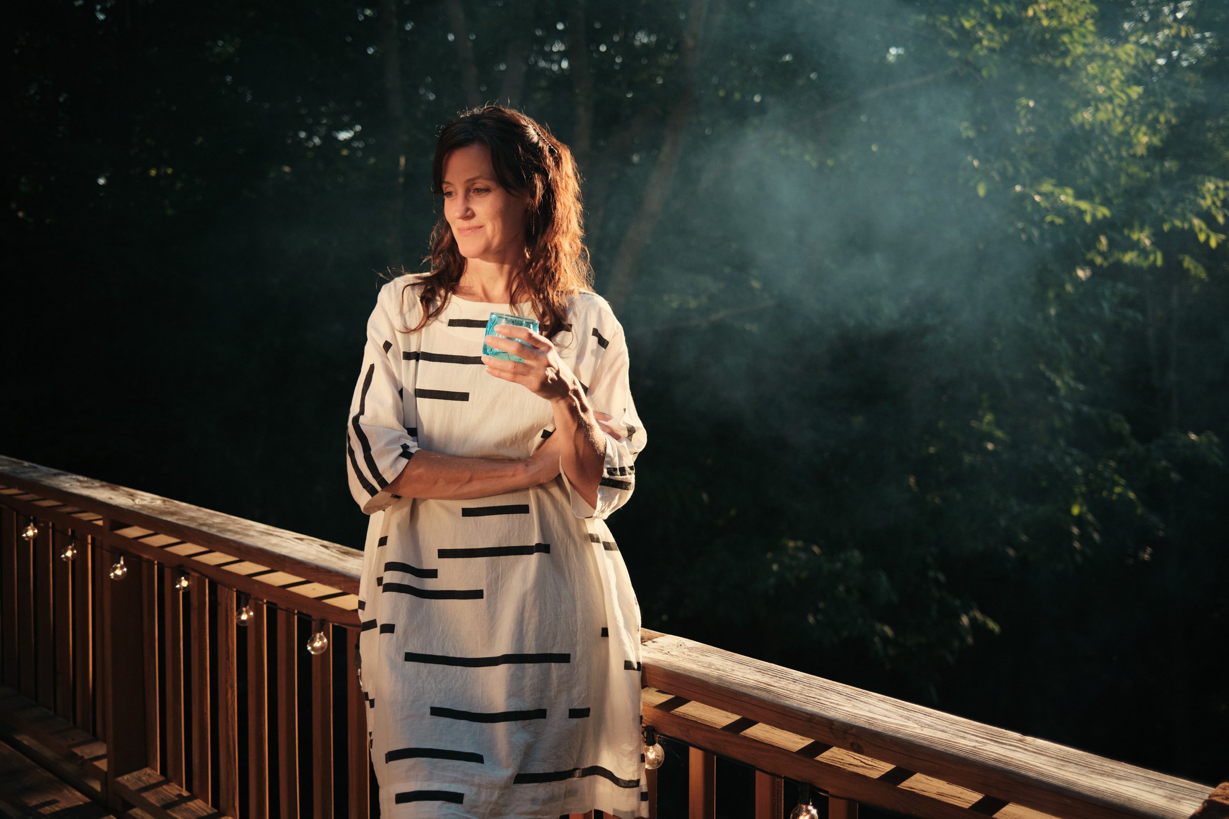 A woman with dark brown hair wearing a white dress with black horizontal stripes, holding a glass of blue drink, standing on a wooden deck with string lights, surrounded by a forest with sunlight filtering through trees.