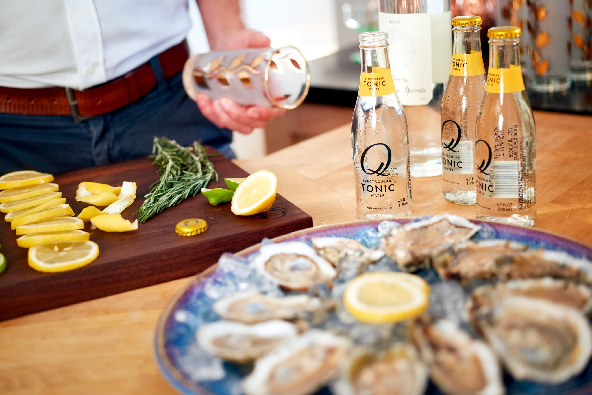 corporate lifestyle photography of a cocktail setup with Q Tonic bottles, sliced lemons, fresh herbs and a tray of oysters on a kitchen counter