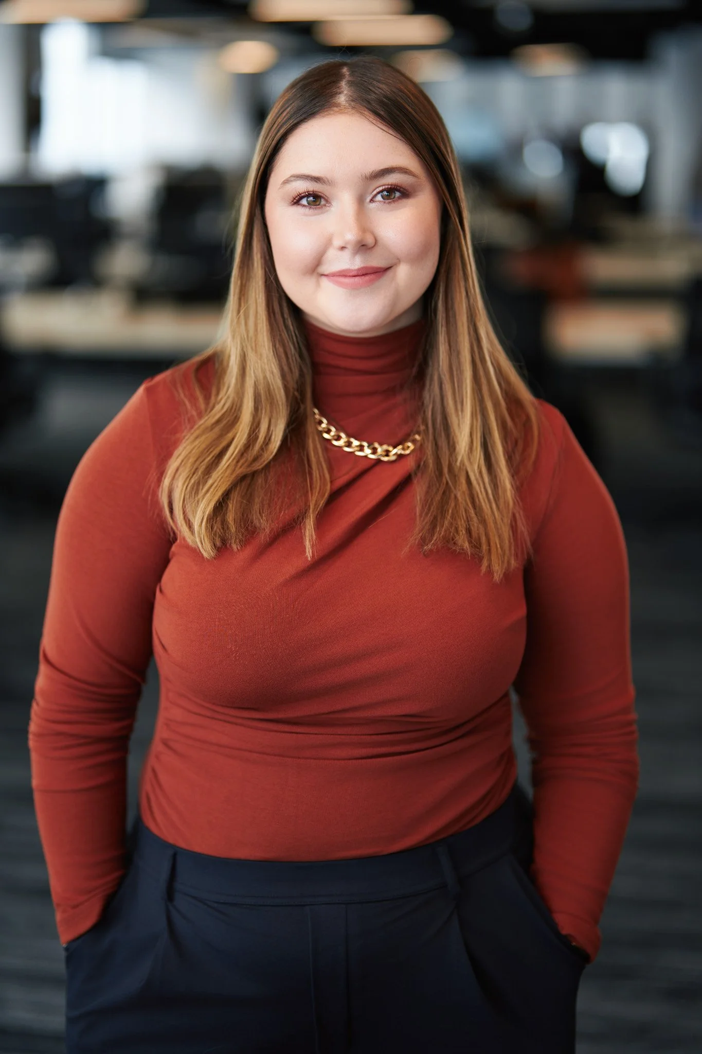 Professional portrait of a woman standing in a modern New York City office, wearing a rust-colored top with a softly blurred background