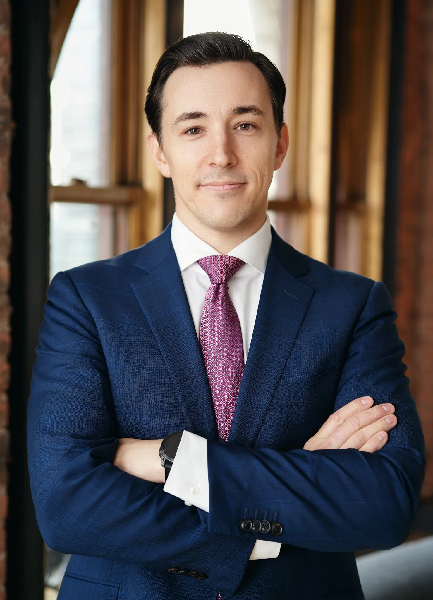 Portrait of a young man in a navy suit standing with arms crossed indoors, photographed in New York City with warm wood window details behind him