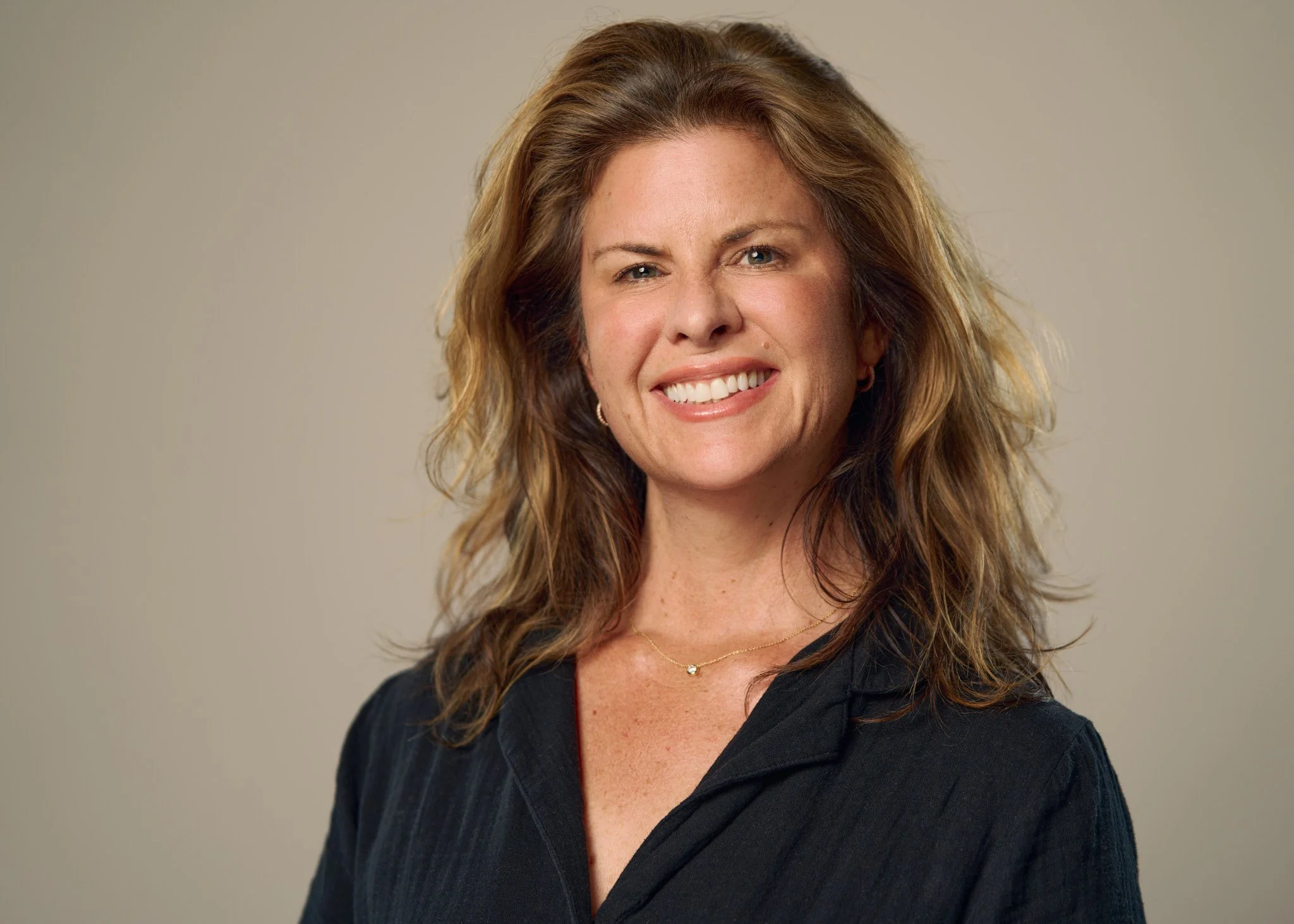 Woman with curly brown hair winking and smiling, wearing a black shirt and a delicate gold necklace, against a plain neutral background.