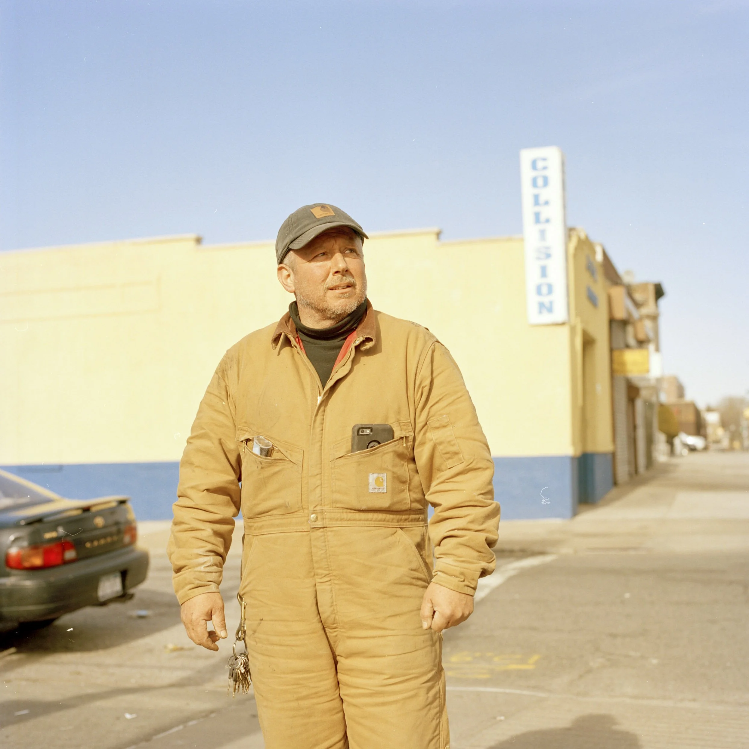 Environmental portrait of a worker wearing tan coveralls and a cap, standing on a city street in warm golden light, shallow depth of field with industrial buildings in the background, photographed in Brooklyn.