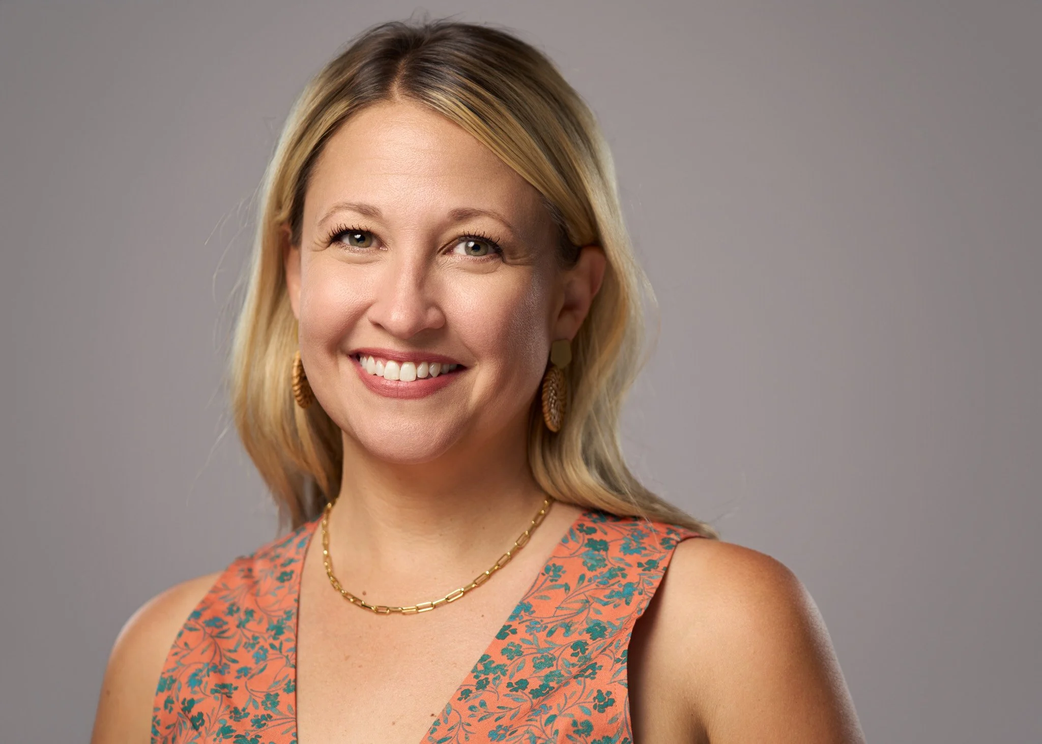 A smiling woman with shoulder-length blonde hair, green eyes, wearing gold earrings, a gold necklace, and an orange sleeveless top with a blue floral pattern, against a plain gray background.