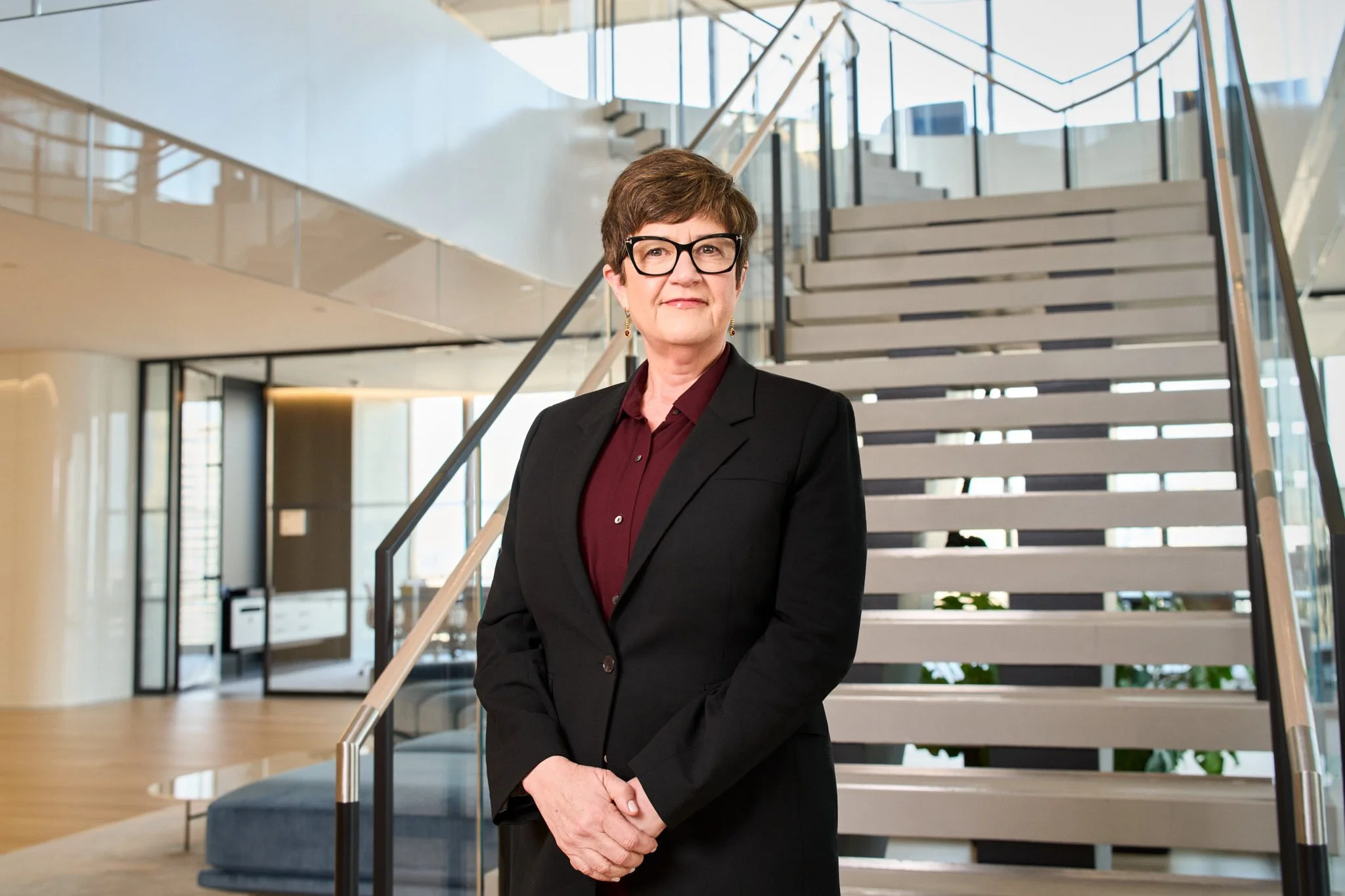 Executive headshot of a woman in a black blazer standing on a staircase in a modern NYC office. A confident, professional corporate headshot NYC style.