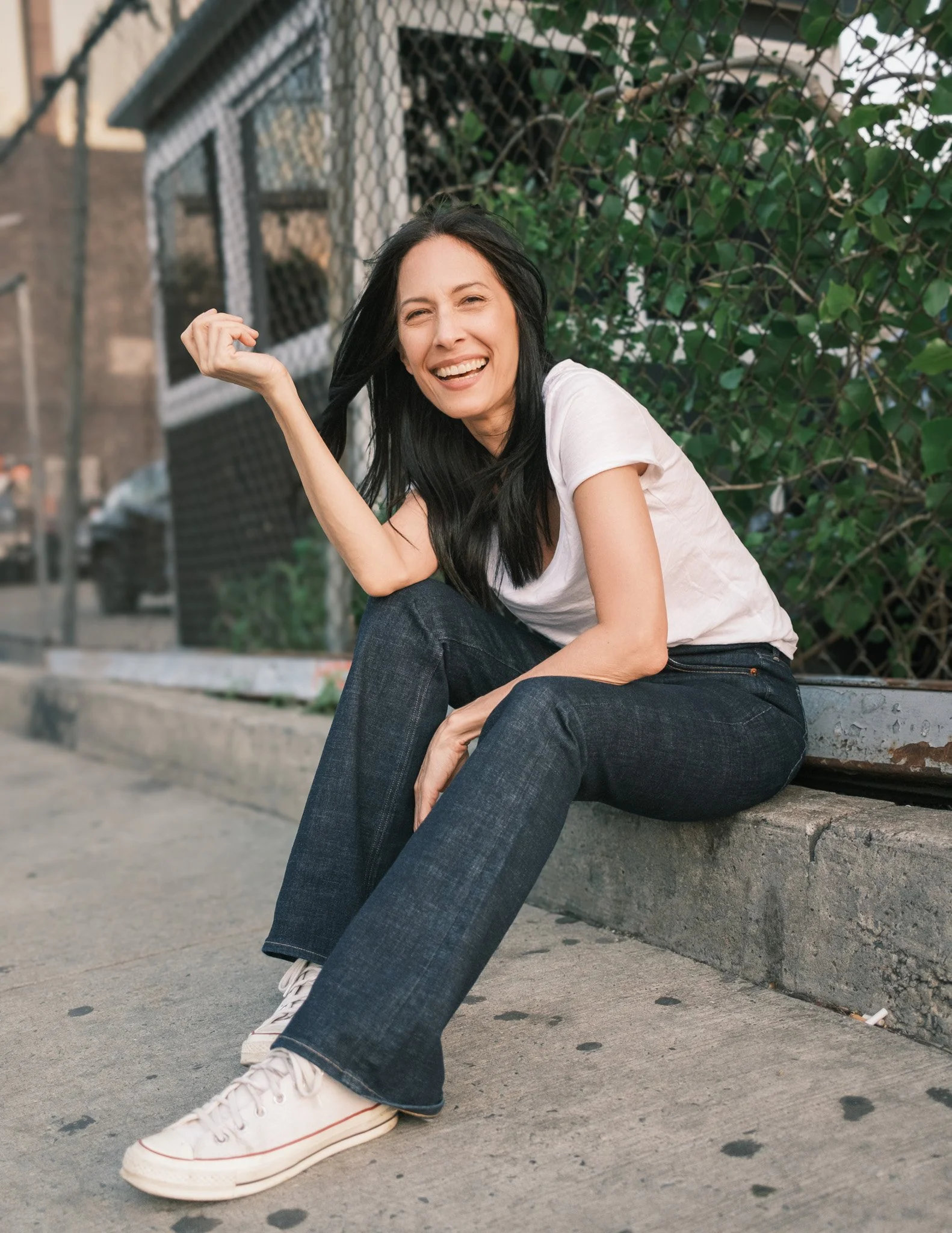 Victoria Gardner creative photoshoot outside sitting on a curb in jeans and white t-shirt wearing white converse sneakers and smiling