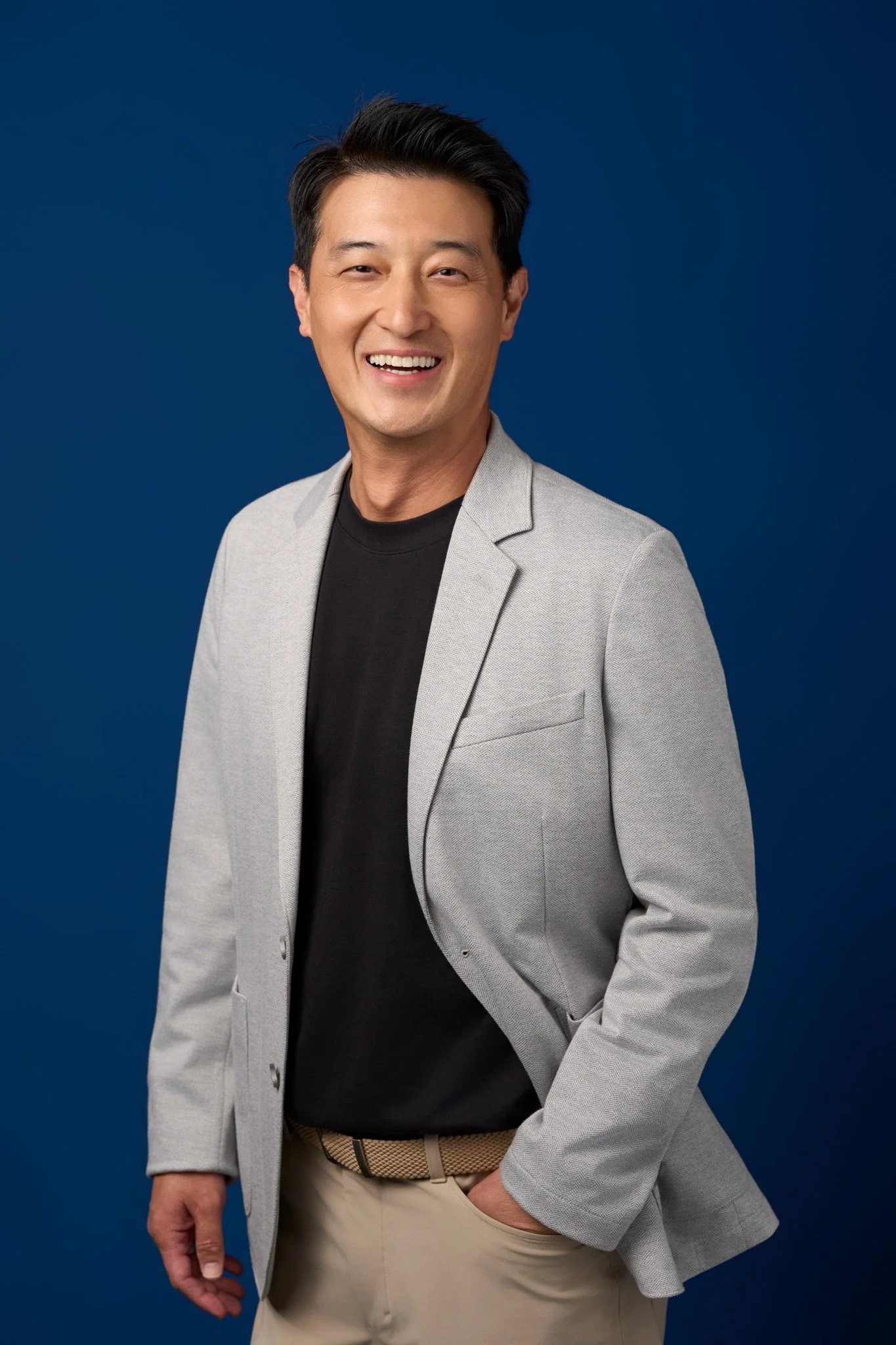Studio portrait of a smiling man wearing a light gray blazer photographed in New York City against a solid blue background