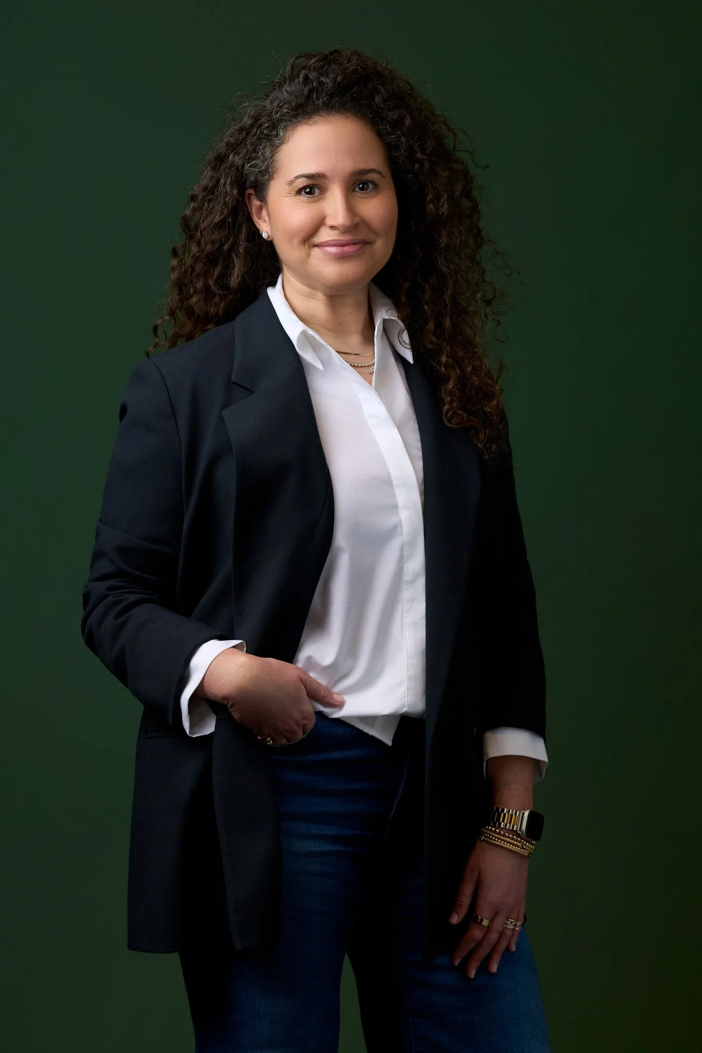 Executive portrait of a woman in a navy blazer and white shirt standing against a deep green studio background, confident stance with soft directional lighting.