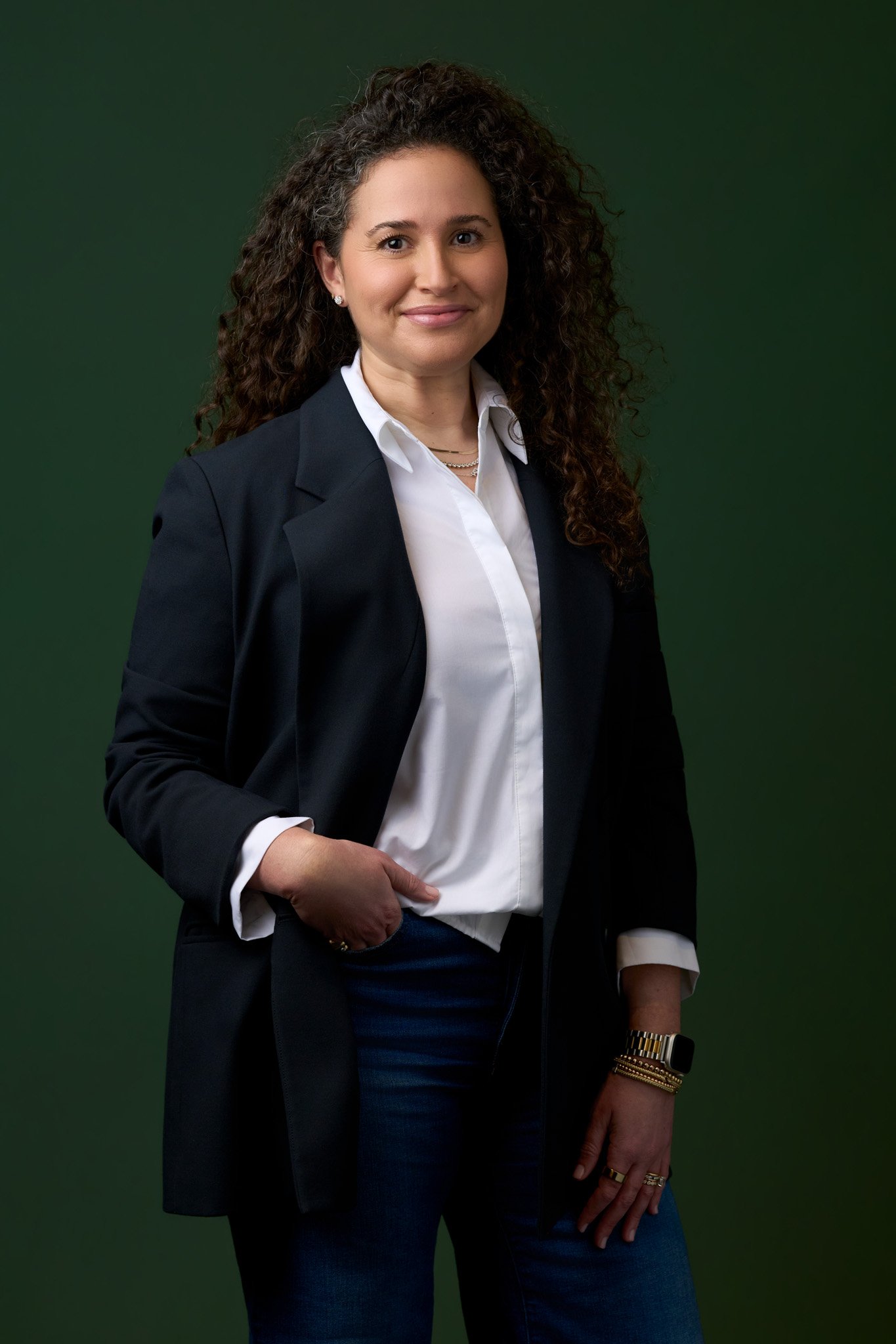 Executive portrait of a woman in a navy blazer and white shirt standing against a deep green studio background, confident stance with soft directional lighting.