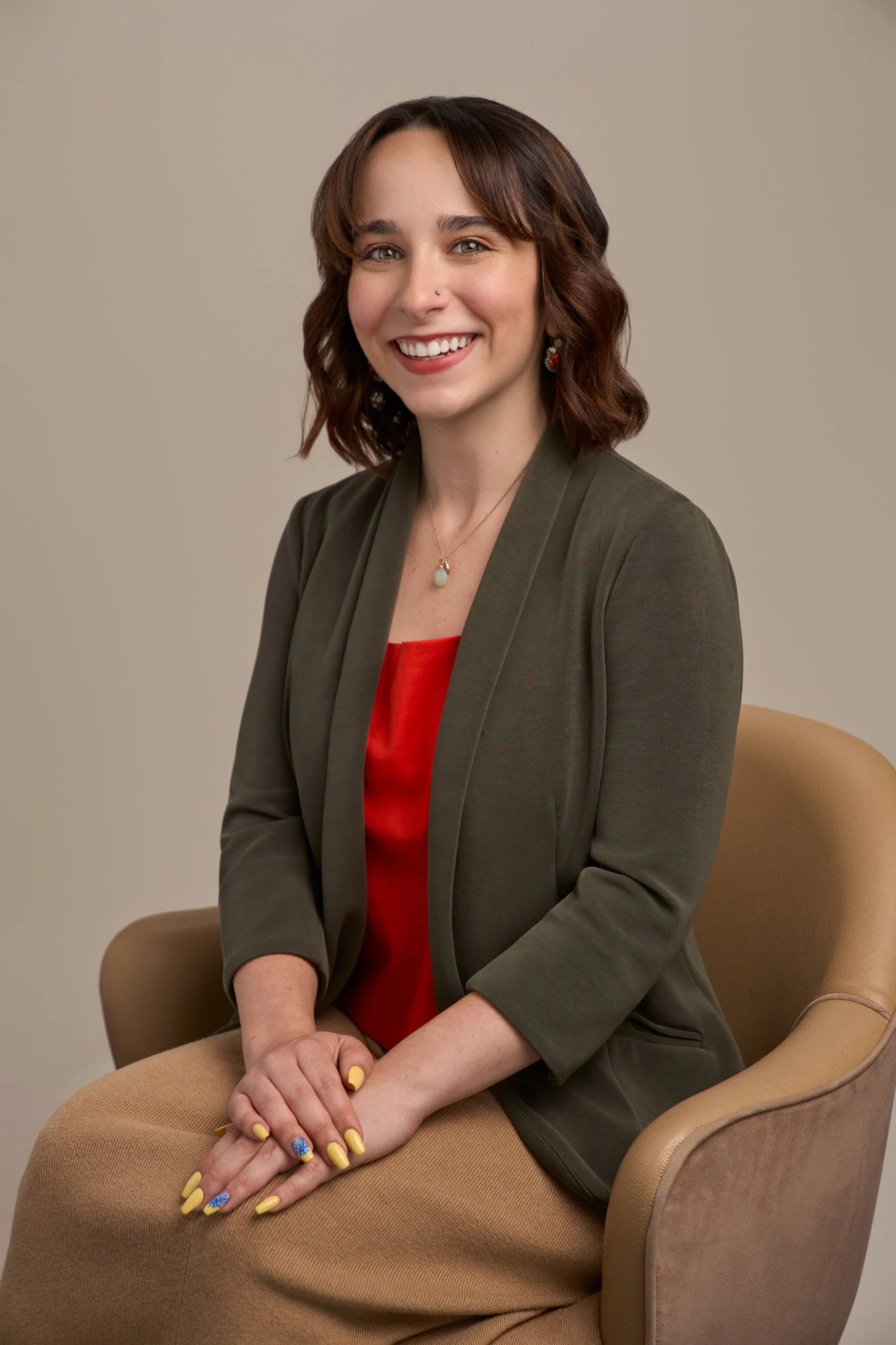Smiling woman in an olive green blazer over a red top seated on tan armchair, professional headshot photographed in New York City