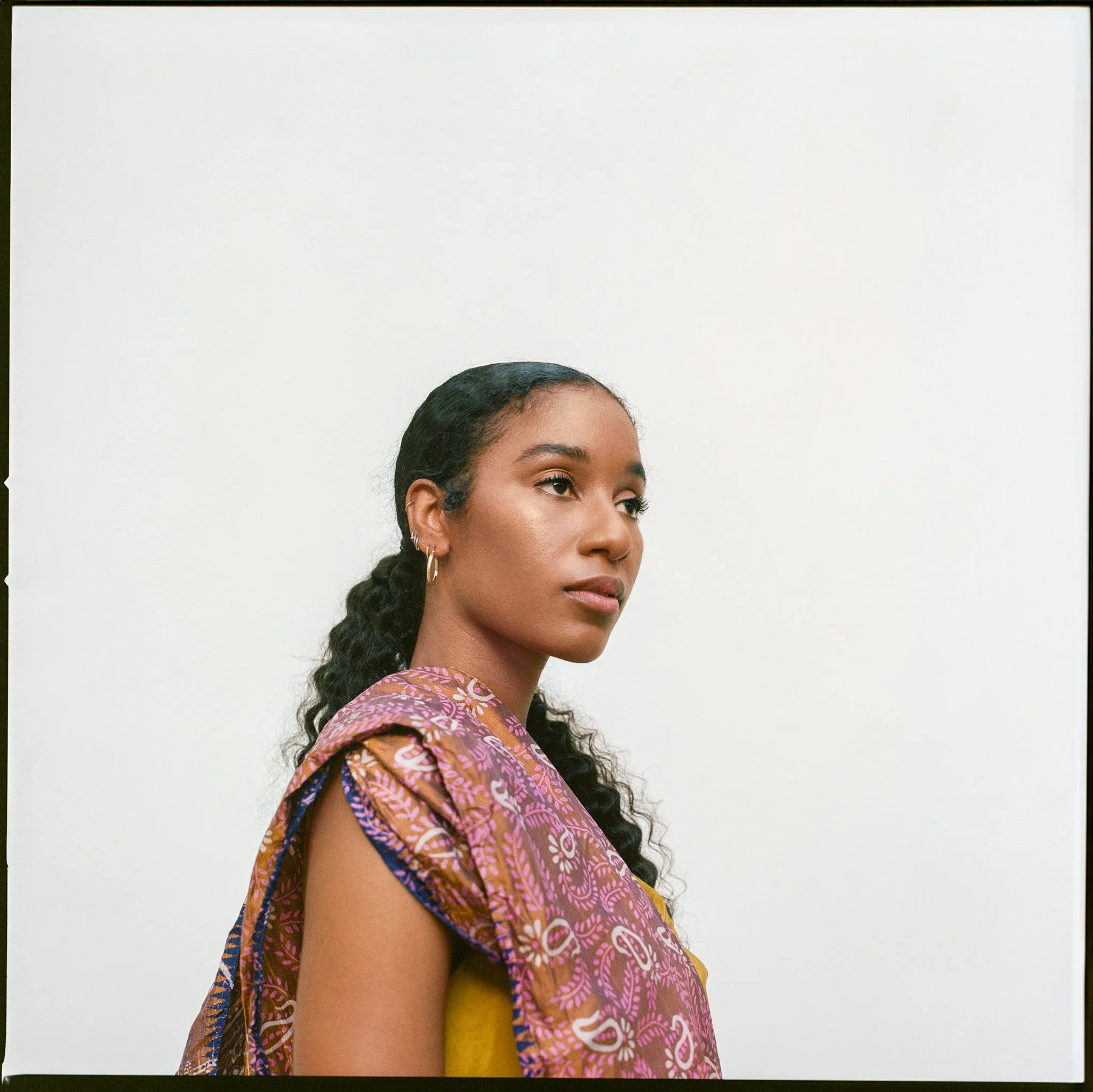 Portrait of a young woman with dark curly hair, wearing gold hoop earrings, and a colorful patterned shawl, standing against a plain white background.