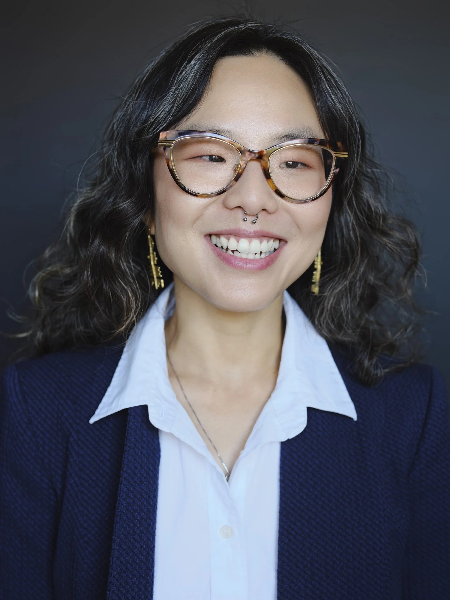 Professional headshot of a woman with curly hair and glasses, photographed in New York City against a dark studio background