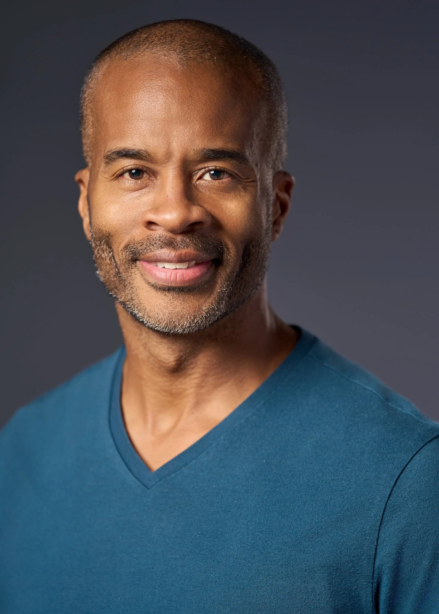 Actor headshot of a man with closely cropped hair and light stubble, relaxed and approachable, wearing a teal V-neck shirt, photographed against a dark gray studio background by a NYC headshot photographer, David Pexton Photography