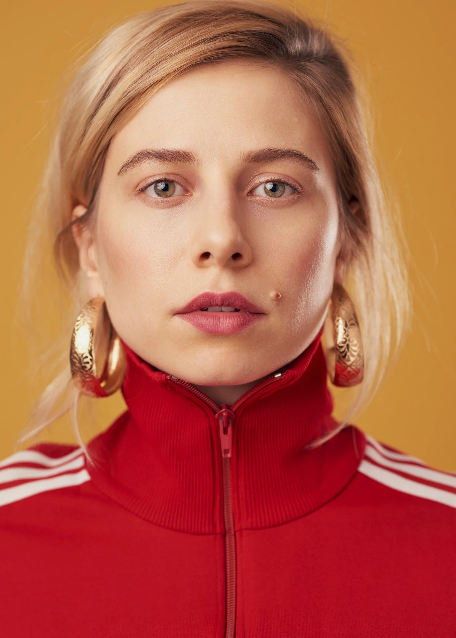 Actor headshot of Victoria Ratermanis against a bold yellow background, wearing a red zip top and large gold earrings, direct gaze with clean studio lighting, photographed in New York City.