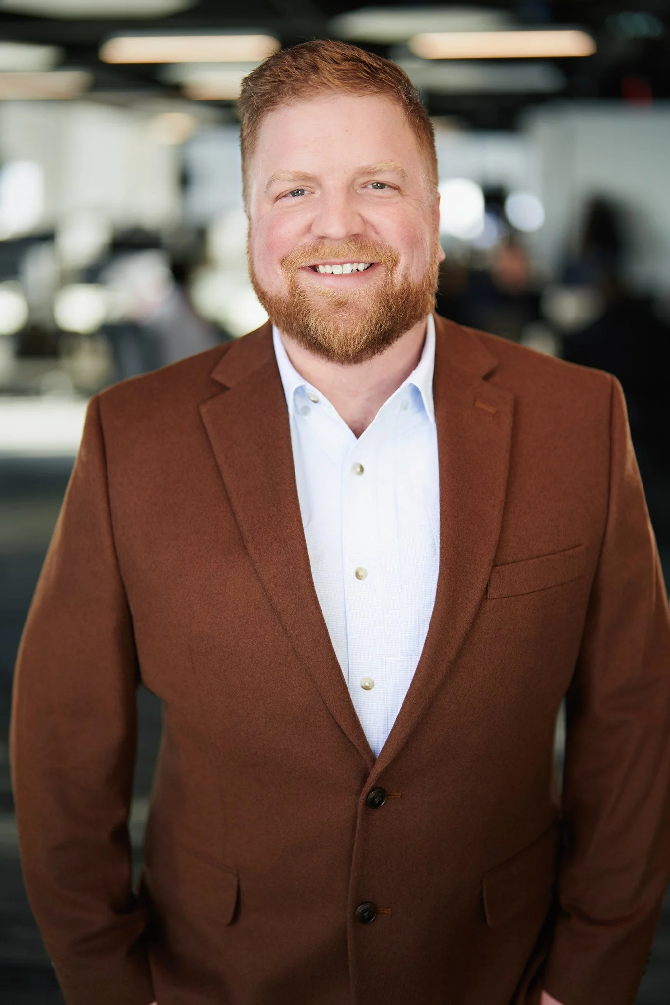 Portrait of a smiling man with red hair and beard, wearing a brown blazer and white shirt, in an indoor office setting.