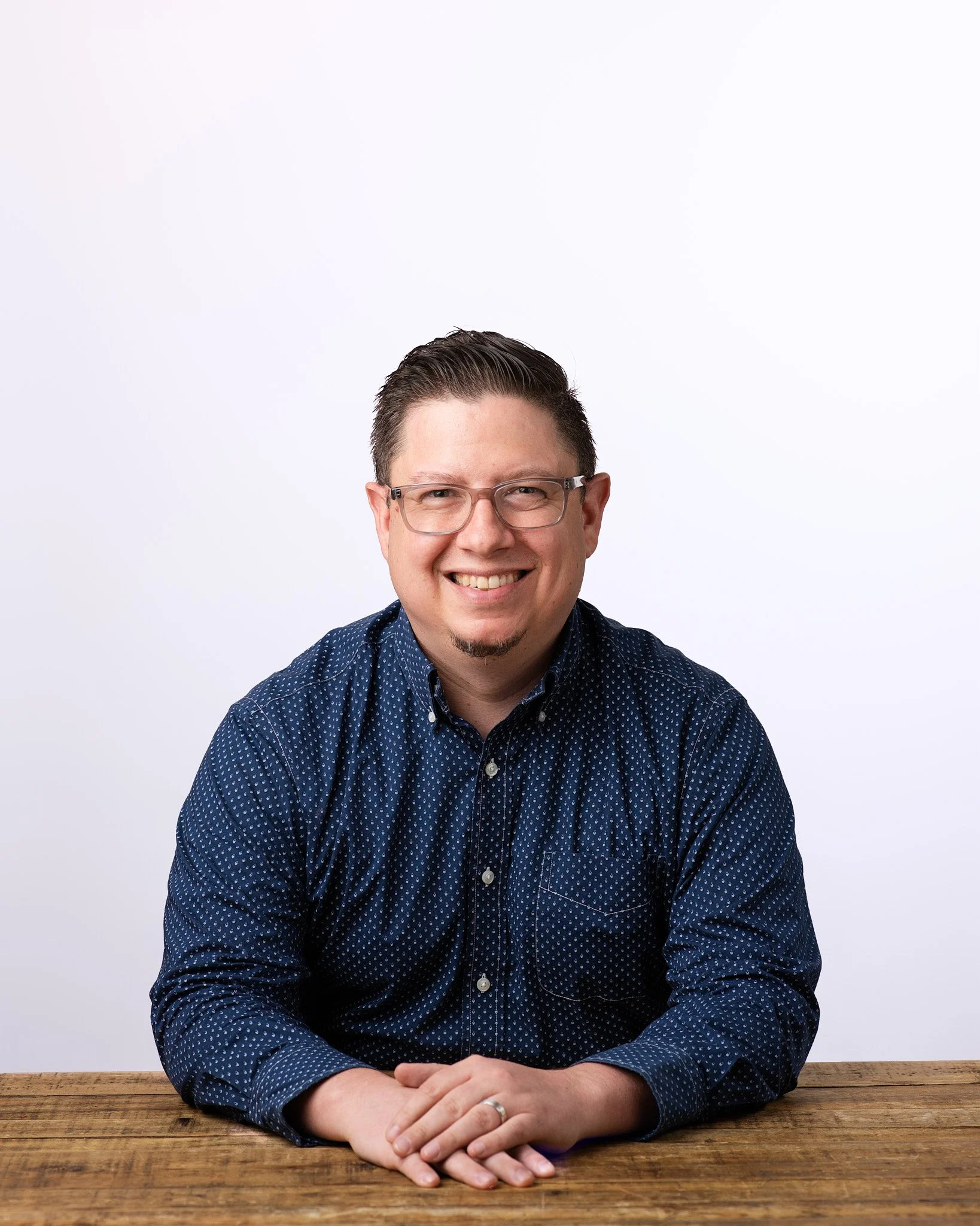 professional headshots nyc of a man in glasses and a blue patterned shirt seated at a wooden table with clean even lighting