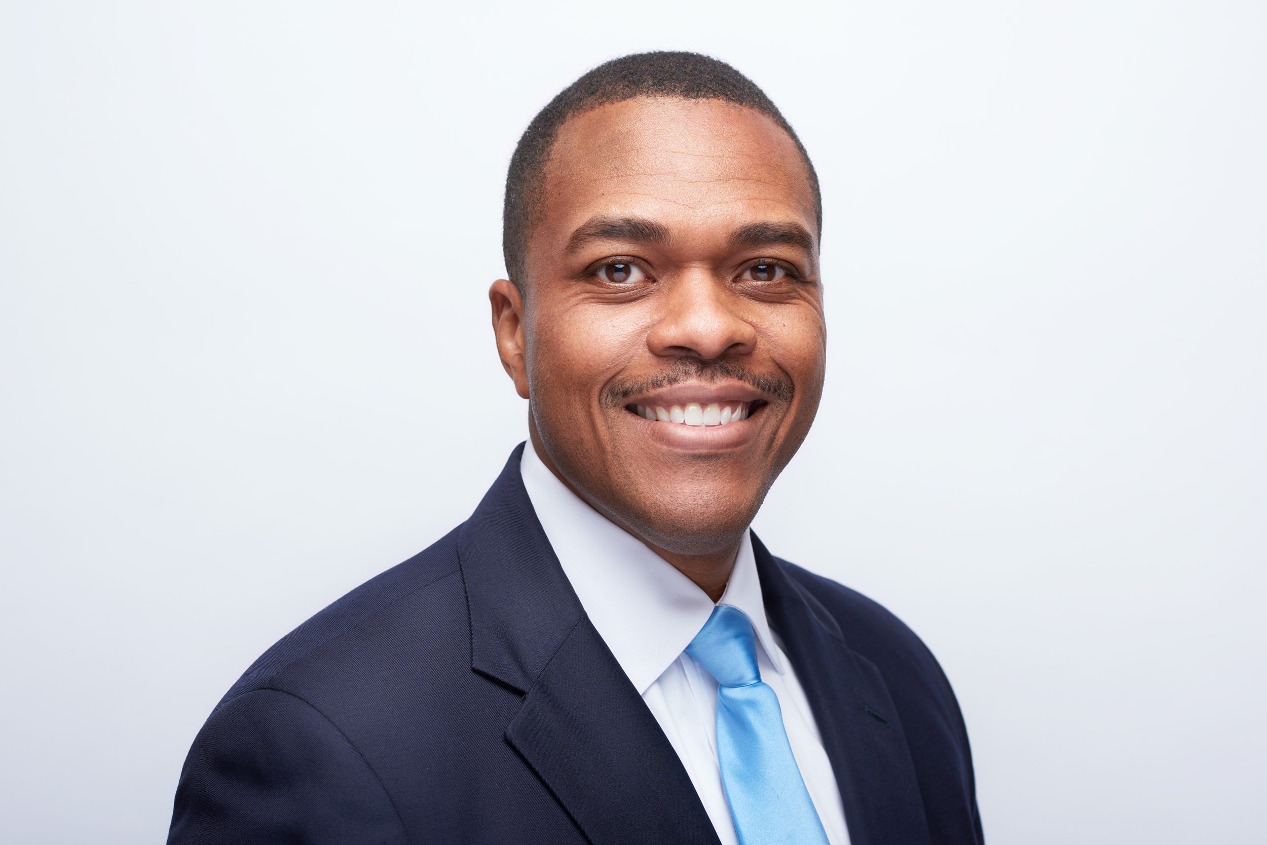 Professional African American man in a navy suit, white shirt, and light blue tie smiling against a plain white background.