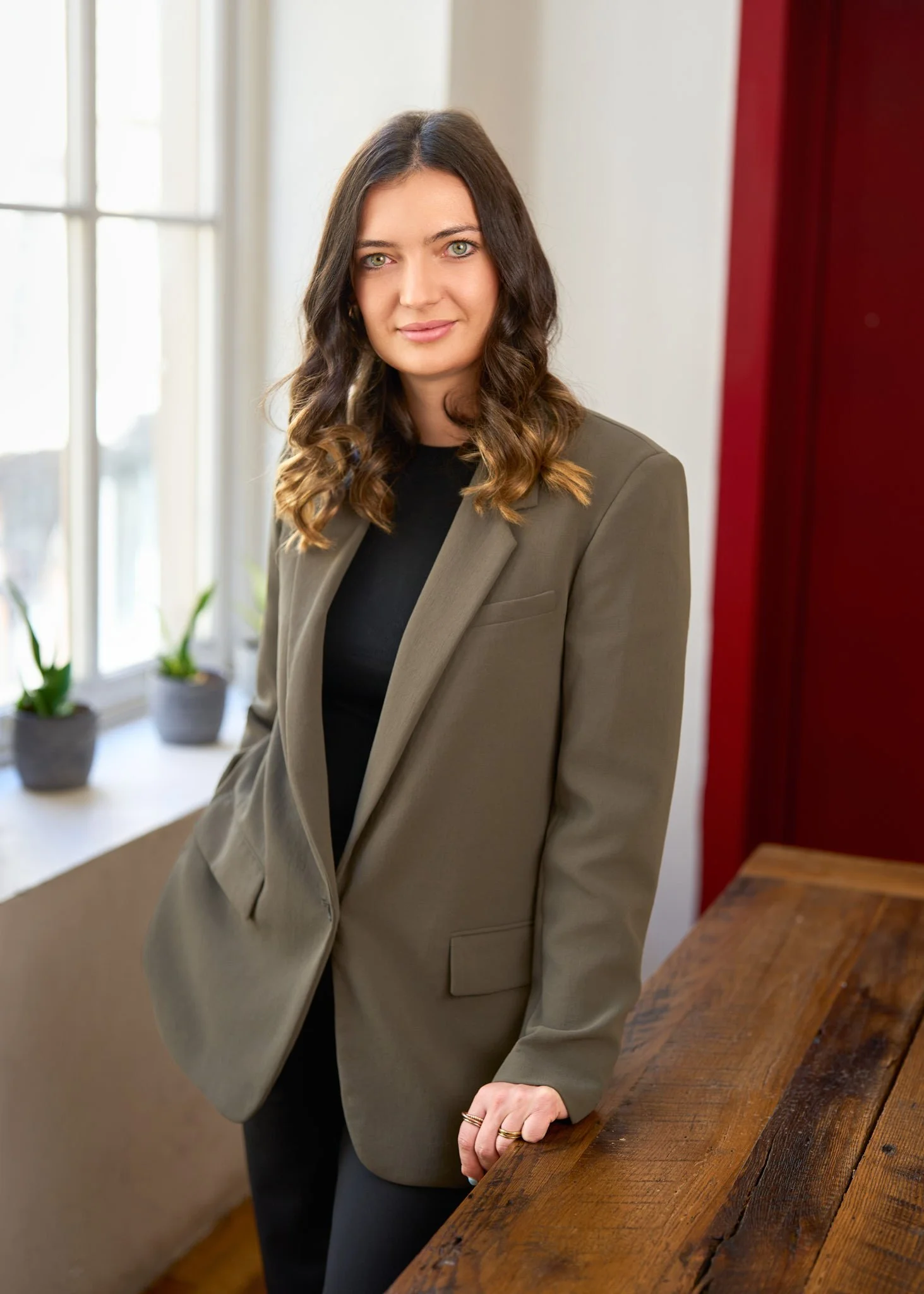 A woman with shoulder-length brown hair and green eyes, wearing a black top and an olive green blazer, standing inside next to a wooden table, with plants on a windowsill behind her.