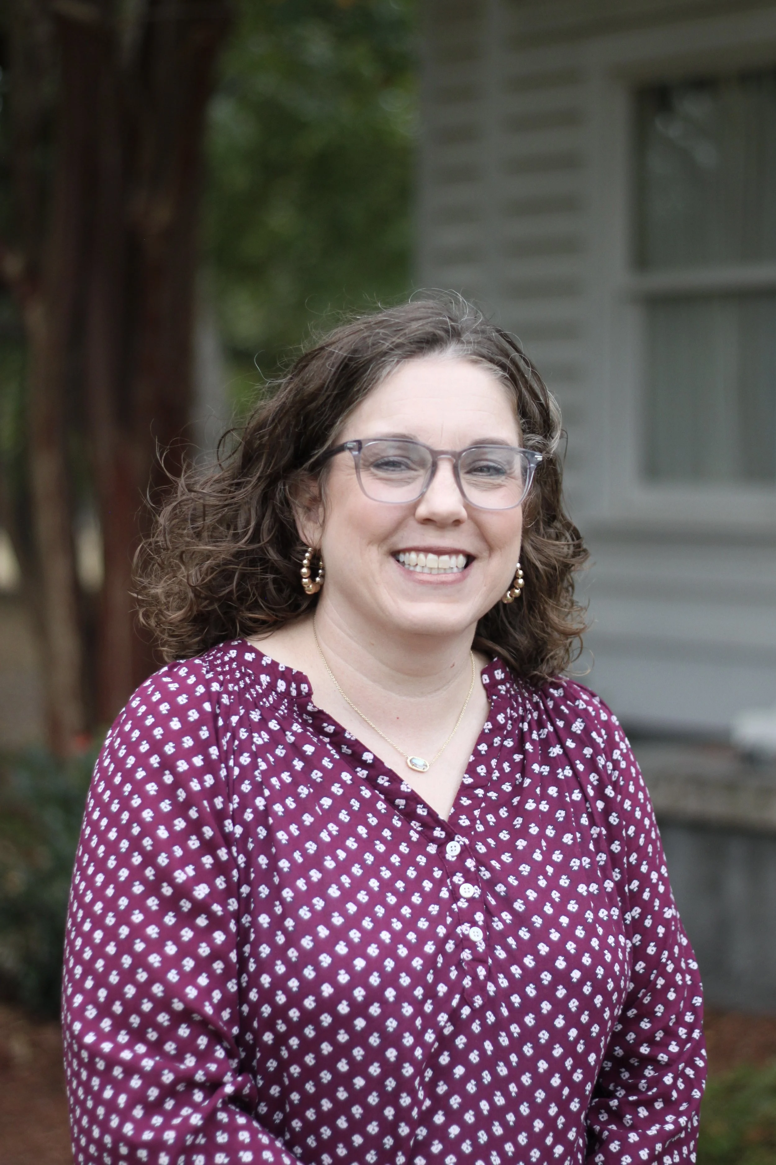 A woman with curly brown hair, glasses, and pearl earrings smiling outdoors in front of a house with a tree and greenery.