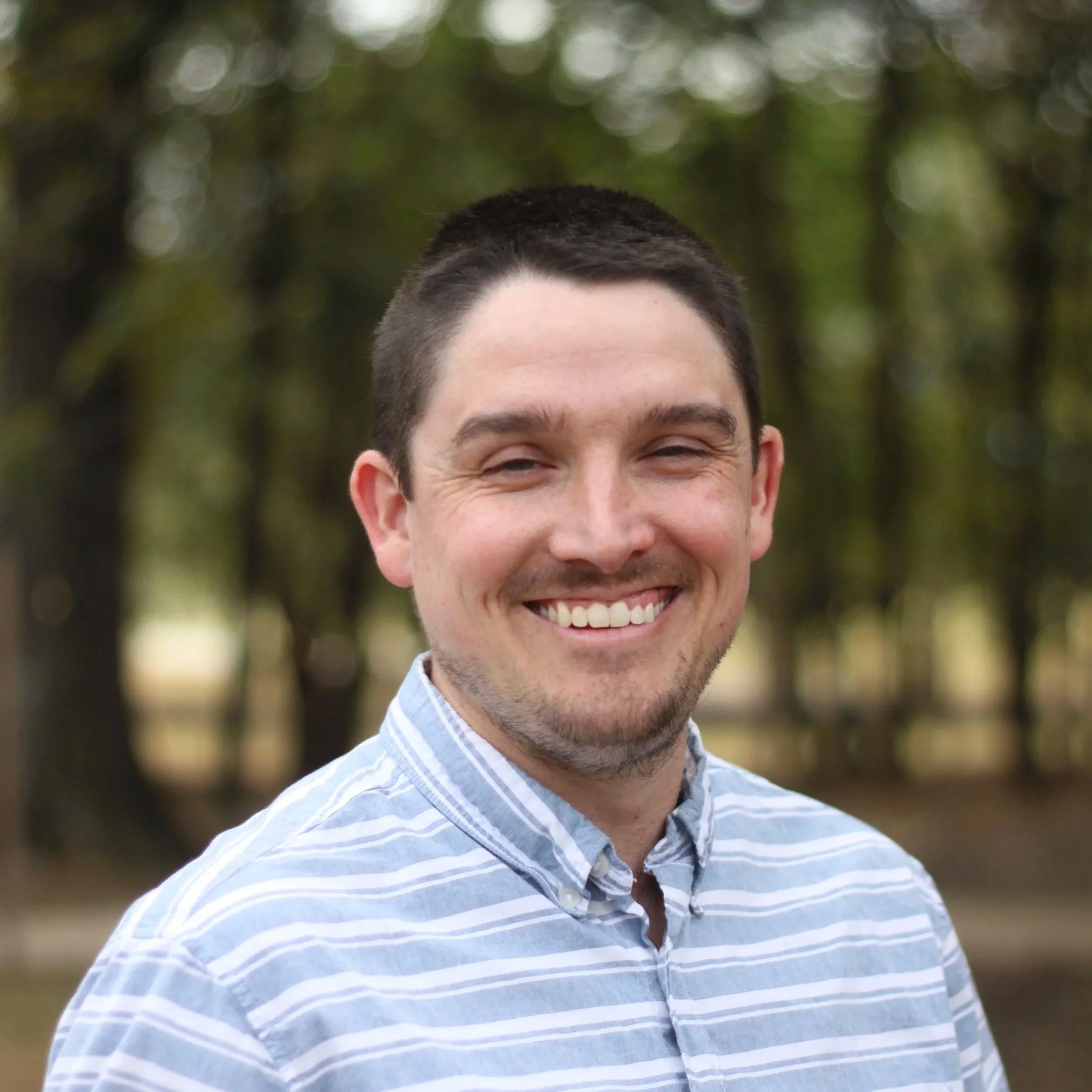 Young man with short dark hair and a beard, smiling, wearing a checkered shirt, standing in front of a white wooden wall.
