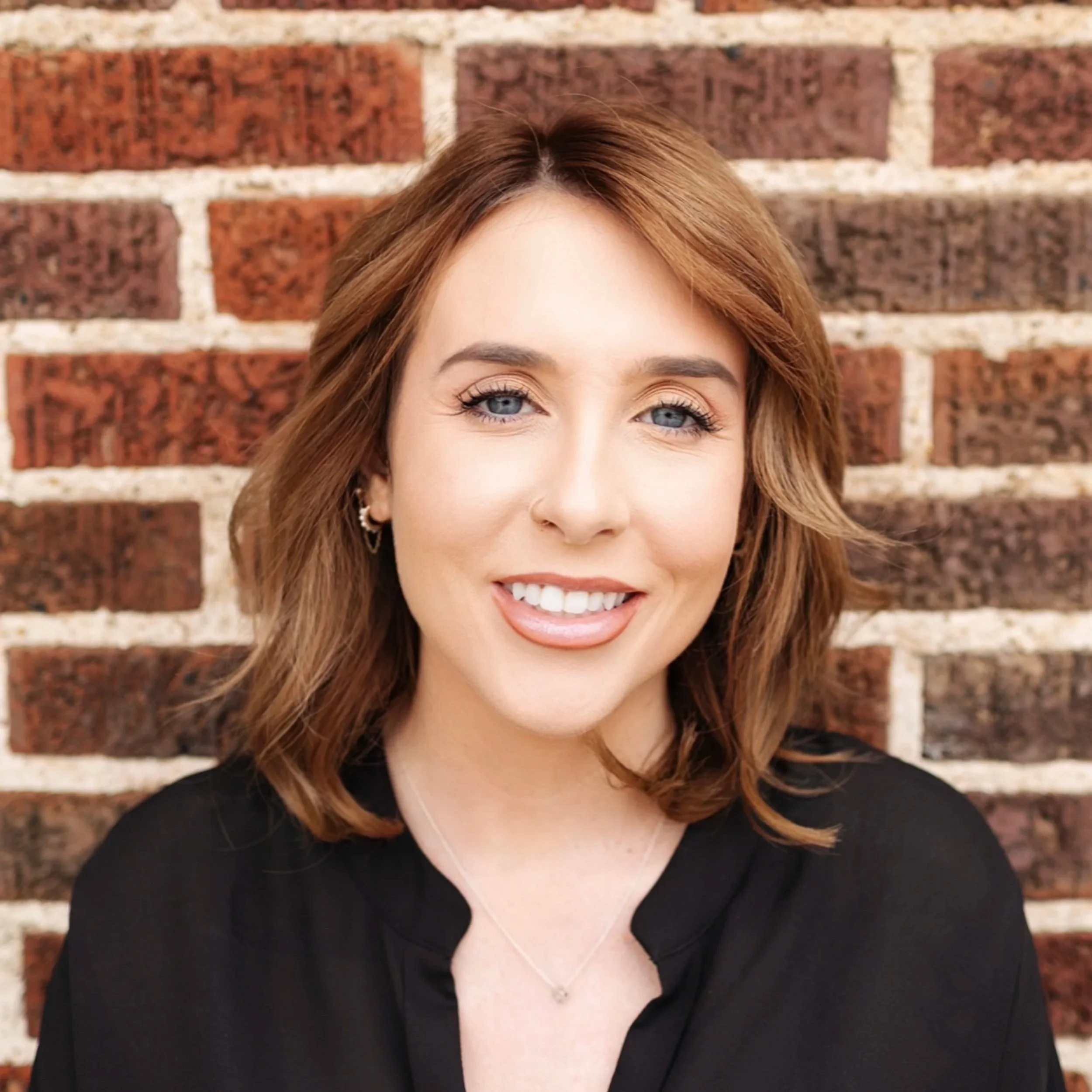 Close-up of a woman's face with light blue eyes, red hair, smiling in front of a red brick wall.