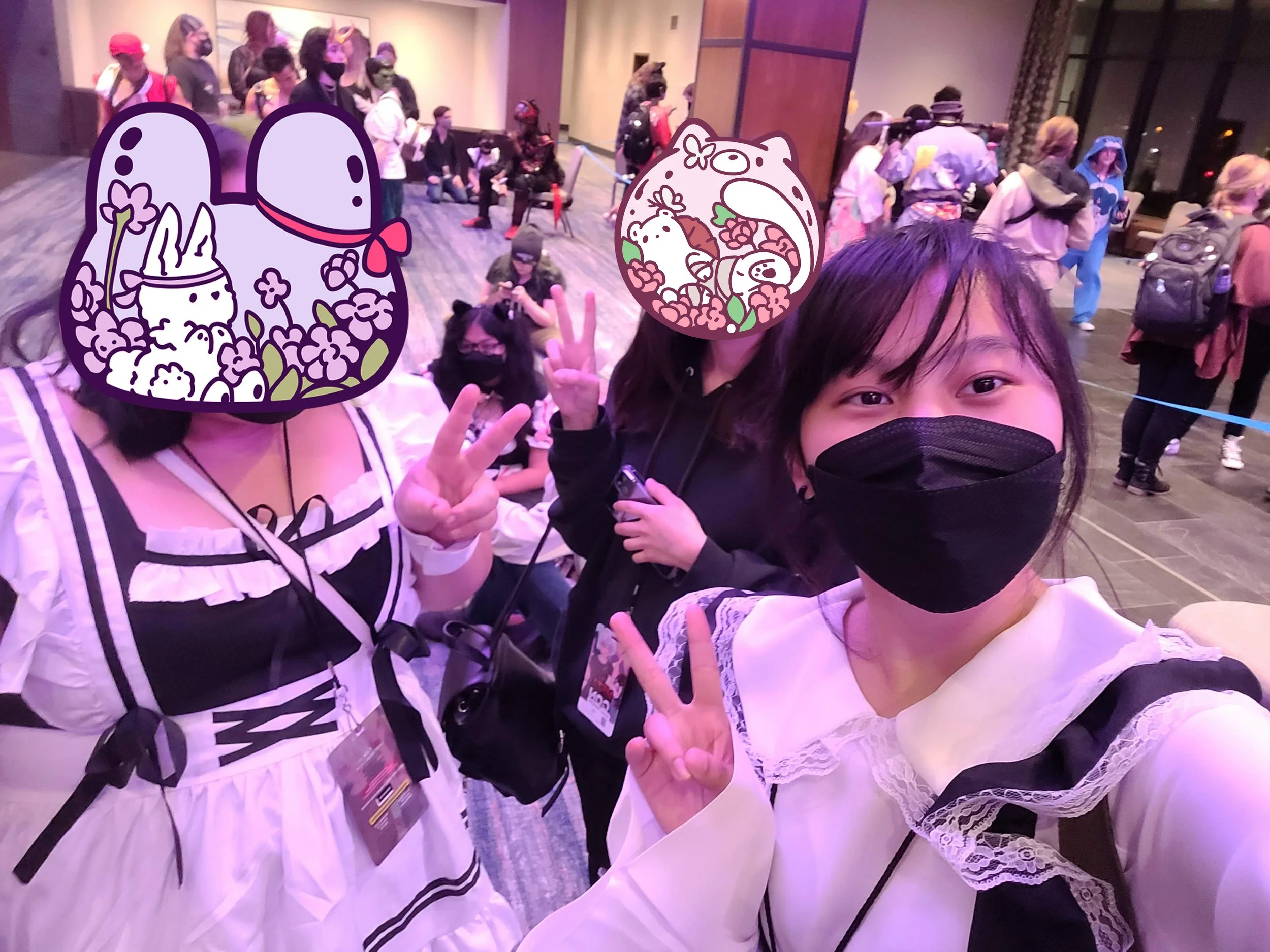 Three young women wearing black face masks taking a selfie at an event, with a crowd of people in the background; the women have cartoon stickers covering their faces, one with a bunny and the other with a bear. All three are making peace signs with their hands.