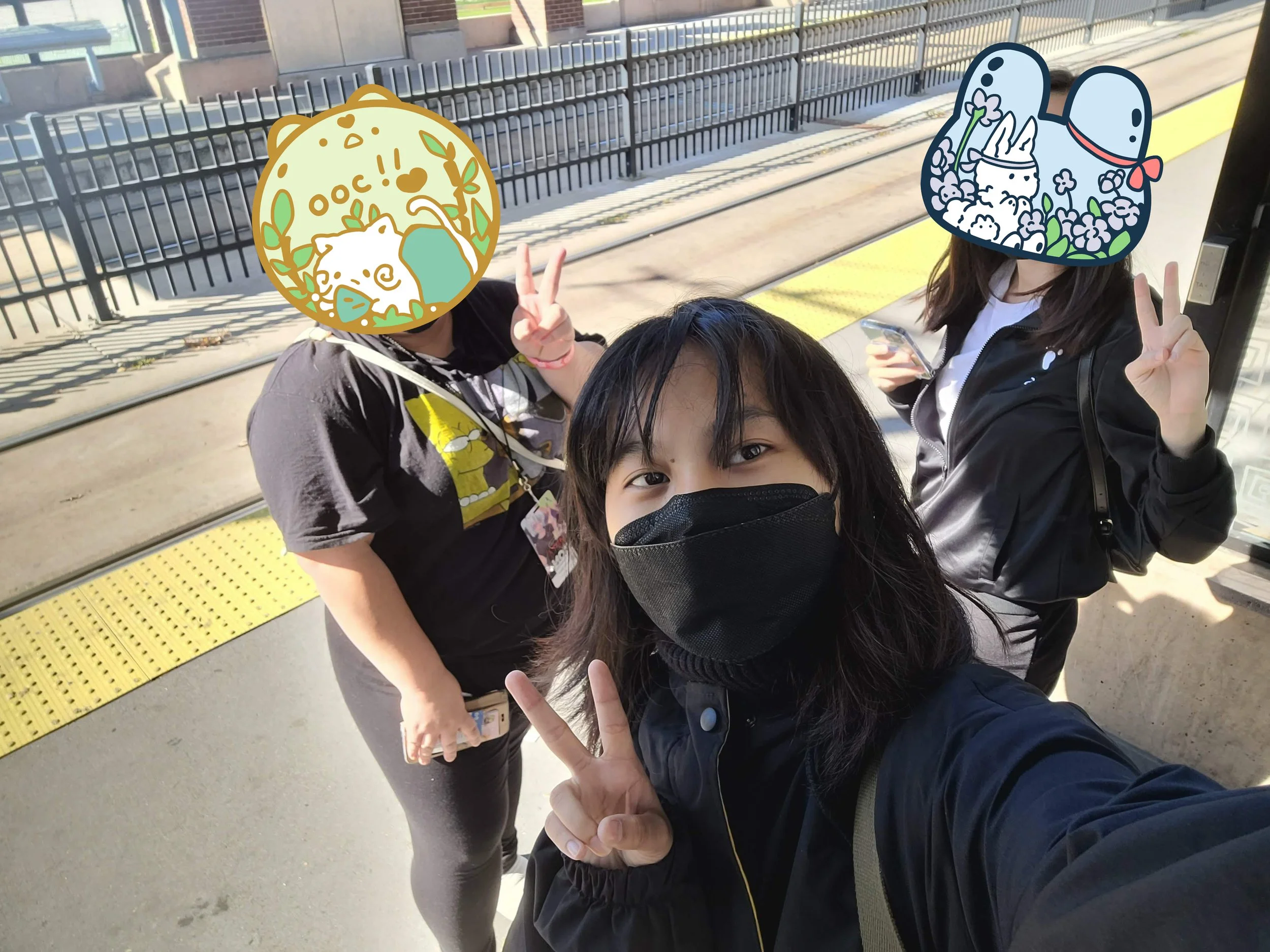 Three women standing on a train platform taking a selfie. All are wearing face masks, and two have cartoon stickers covering their faces, both showing cute animals with peace signs. One woman is holding a phone, and the other two are making peace signs with their fingers.