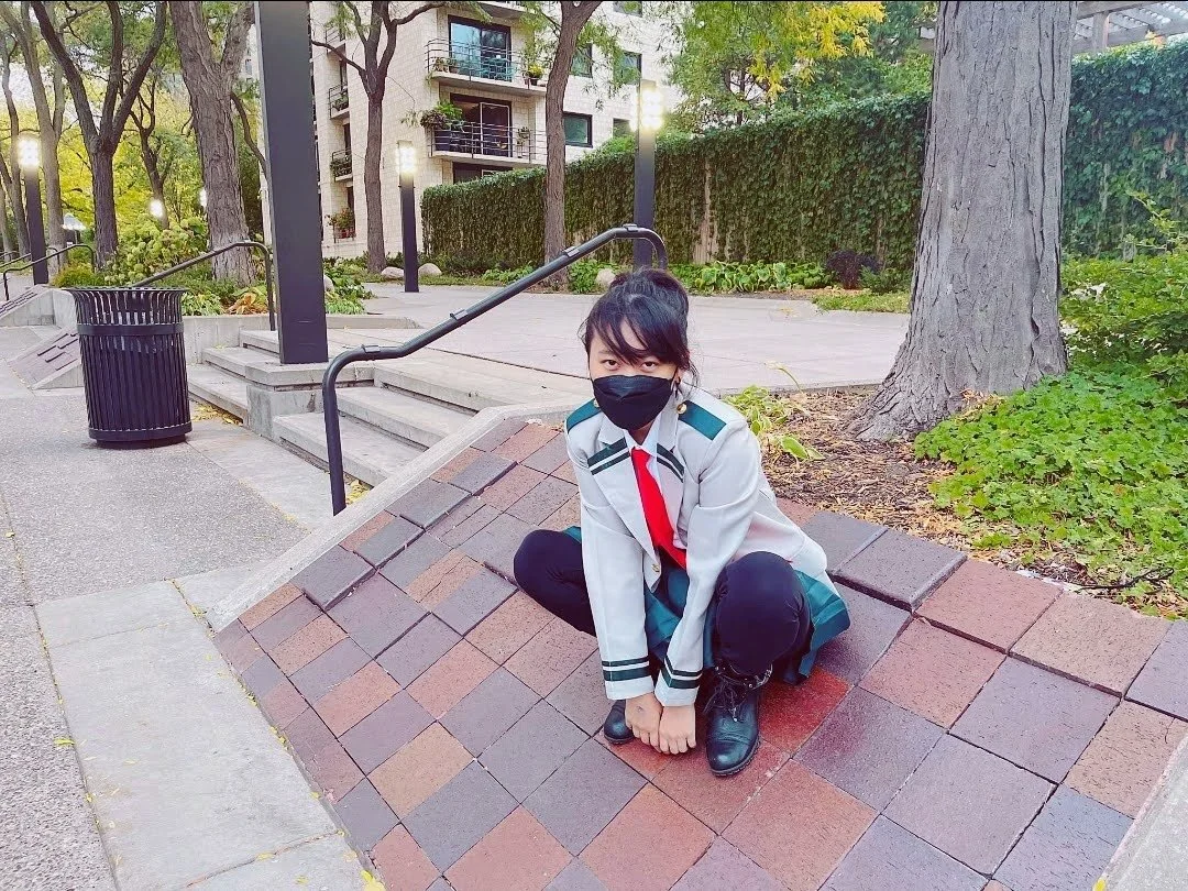 A young woman with dark hair in a ponytail, wearing a black face mask, a gray and green jacket, a white shirt, a red tie, dark pants, and black boots, squatting on a small brick ramp in a park-like setting with trees, bushes, a walkway, stairs, and a building in the background.