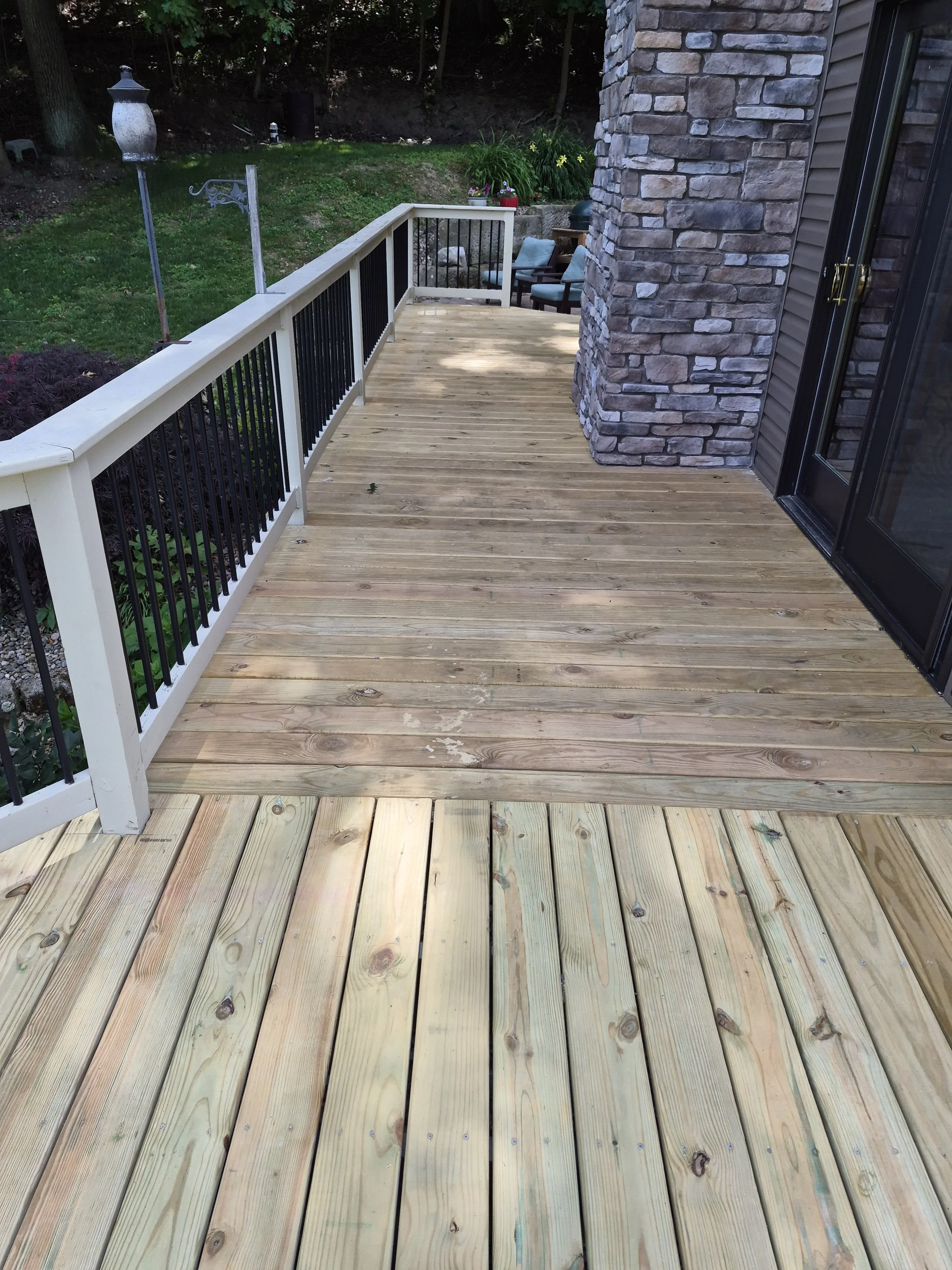 A newly built wooden deck attached to a house with a stone exterior wall and a sliding glass door. The deck has a white and black metal railing and overlooks a green backyard with garden chairs, potted plants, and trees.
