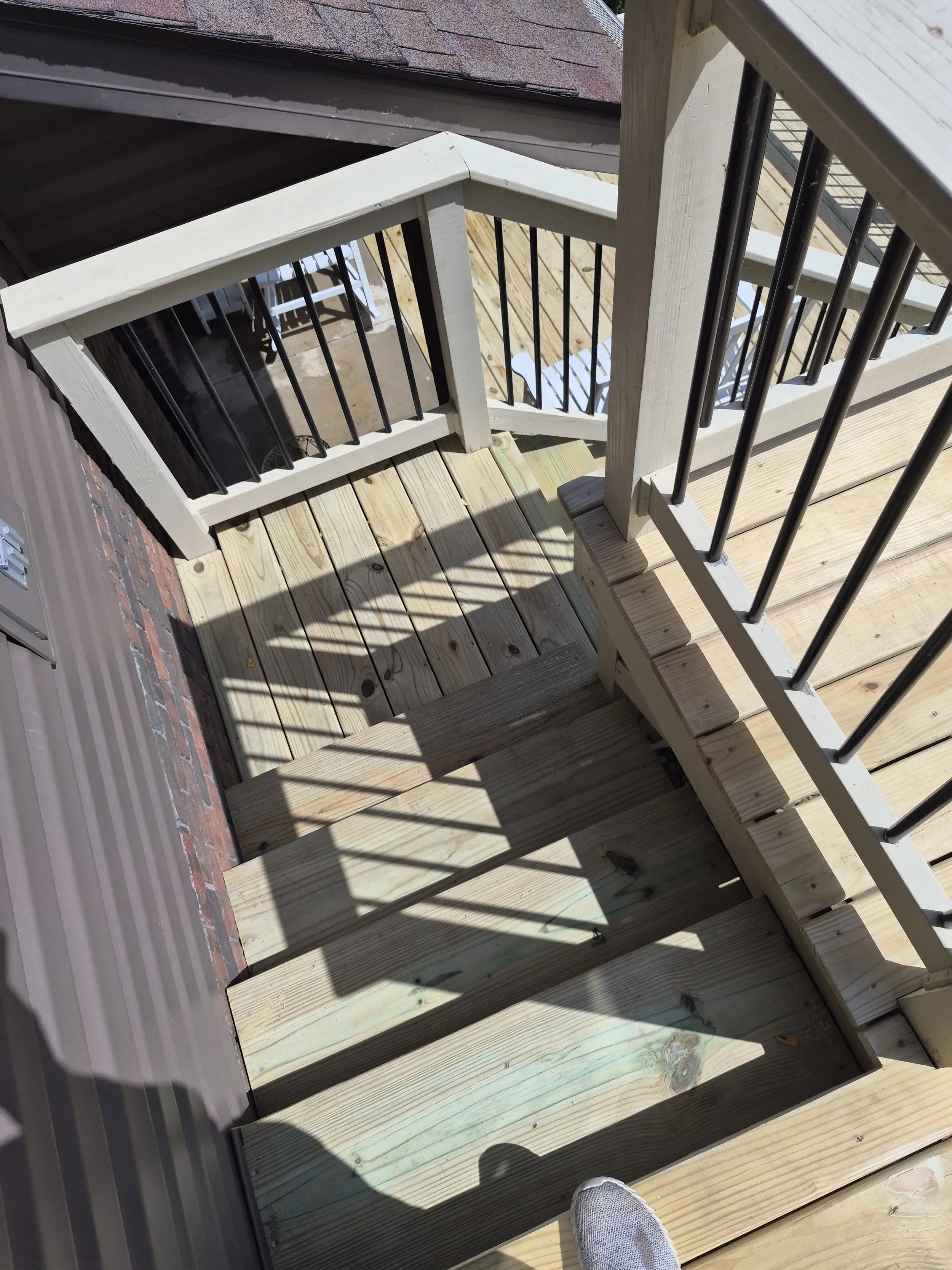 View of a wooden deck with stairs and black metal railings, with a person's foot visible in a sneaker in the bottom corner.