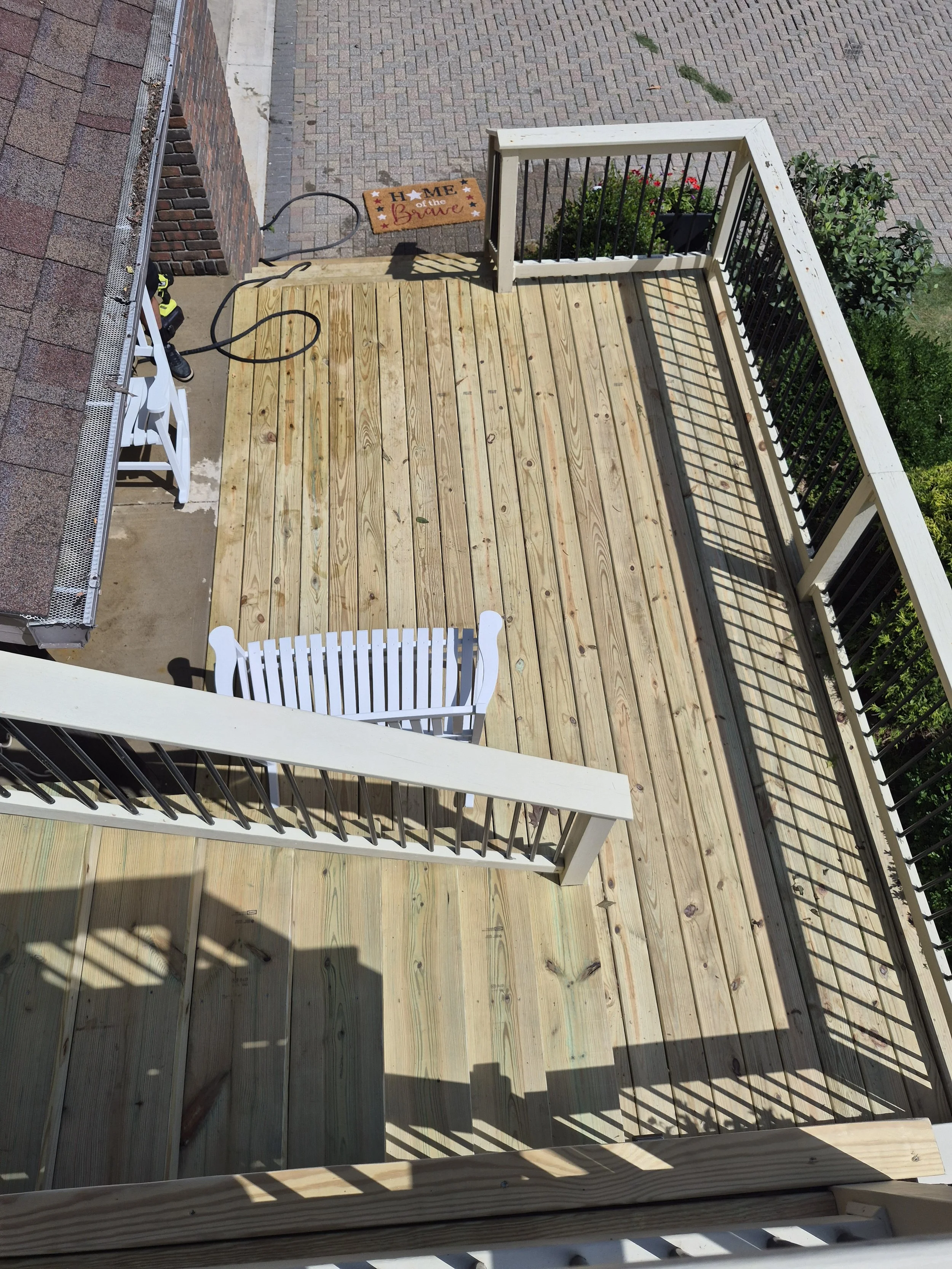A balcony with unfinished wooden flooring, white and black railings, a small white bench, a doormat that says 'Home of the Brave,' and some plants on the side. The balcony overlooks a brick patio and a sidewalk.