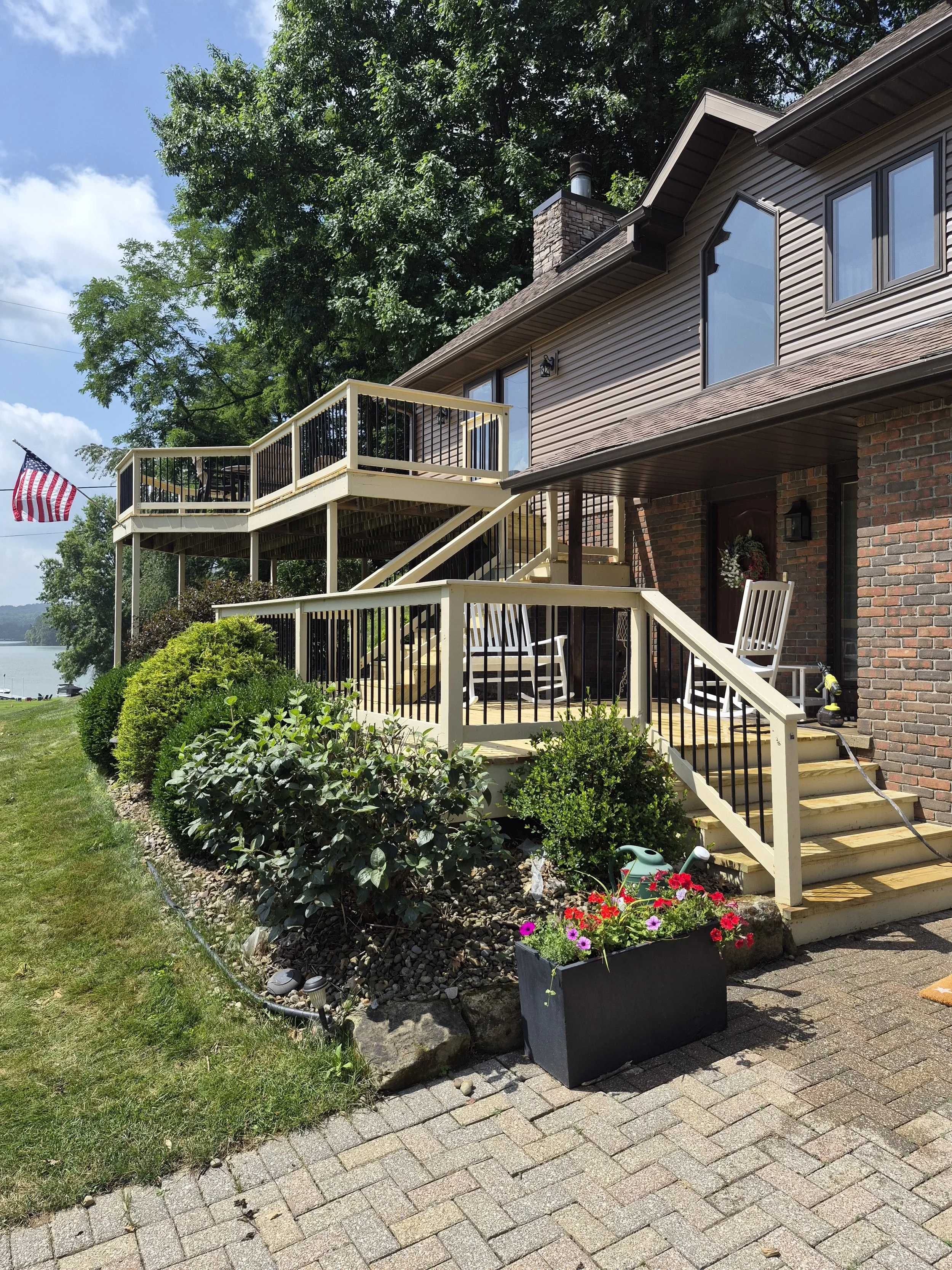 A multi-level brick and siding house with a wooden deck, stairs leading to the yard, and outdoor furniture. There is a flower planter with colorful flowers, shrubs, and a lawn in the front yard. A flag is visible in the background near a body of water, with trees and a partly cloudy sky.