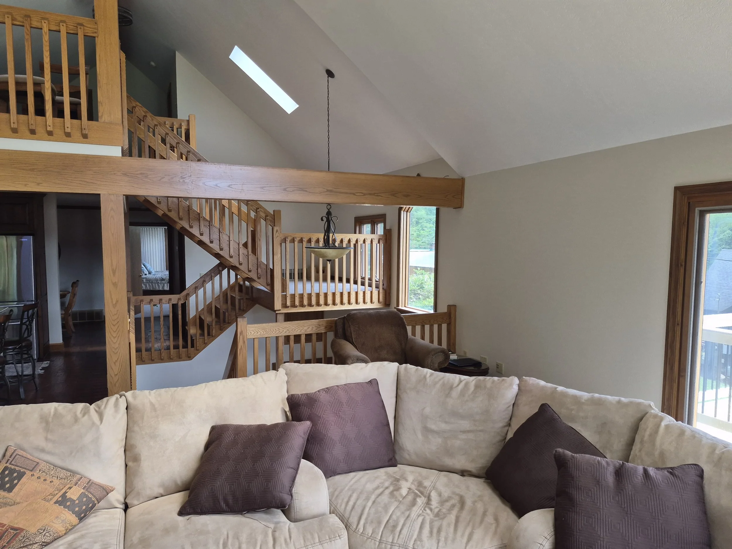 Living room with beige sectional sofa with purple pillows, wooden staircase, and large windows with natural light.