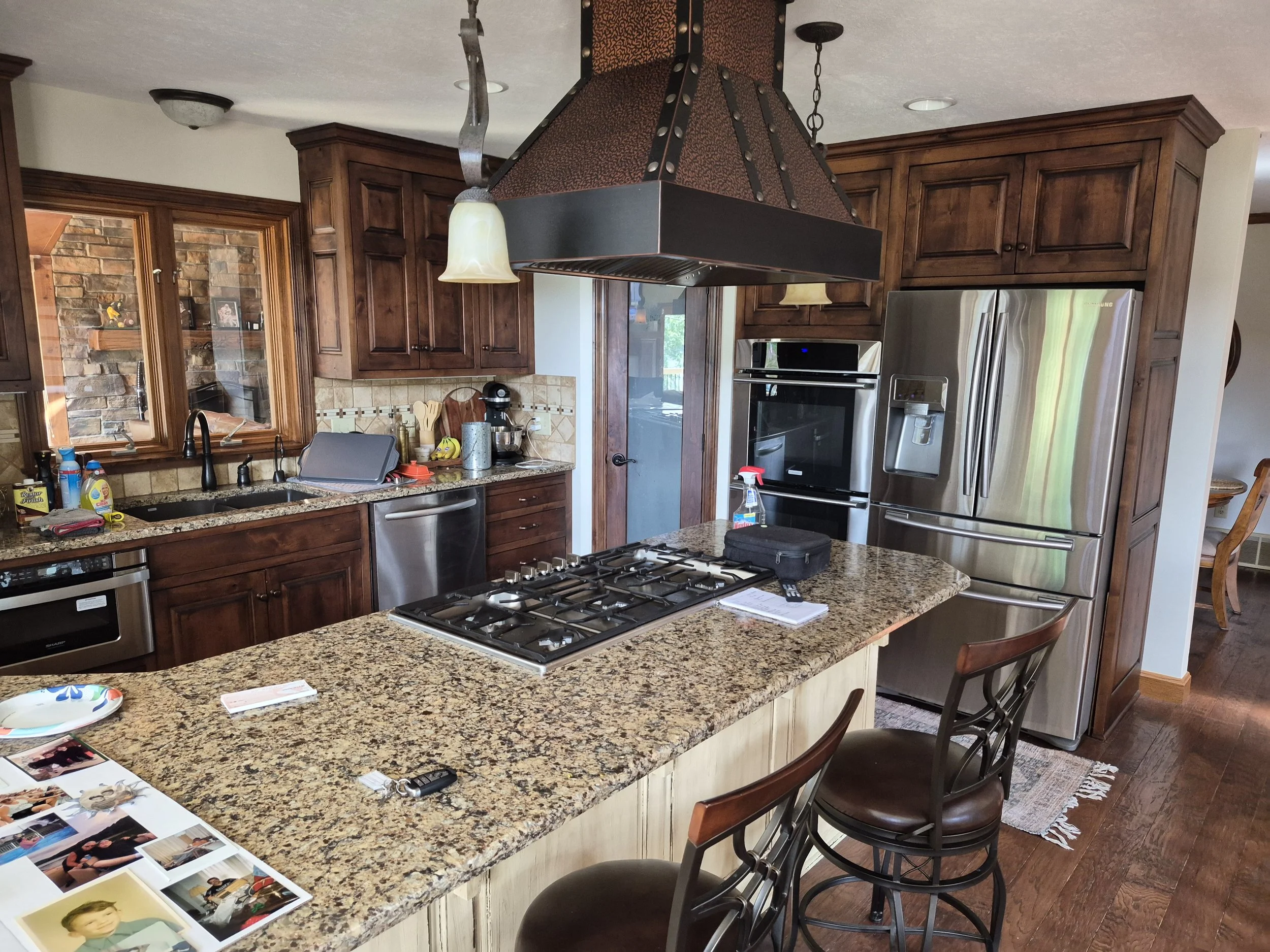 A kitchen with wooden cabinets, granite countertops, and stainless steel appliances. There is a large island with chairs. The kitchen has a range hood above the stovetop, windows, and a variety of kitchen items on the counters. Photos and papers are scattered on the island.