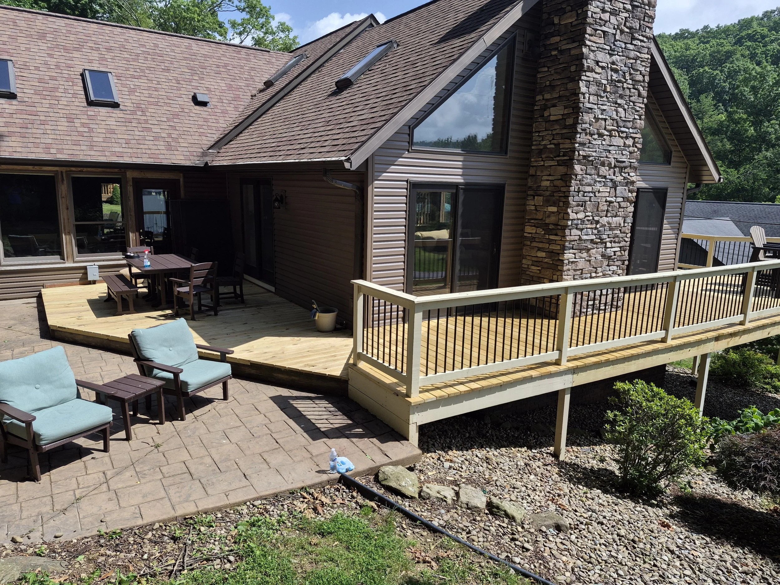 Backyard with a second-story deck, patio, outdoor furniture, a stone chimney, large windows, and a house with brown siding and a shingled roof.