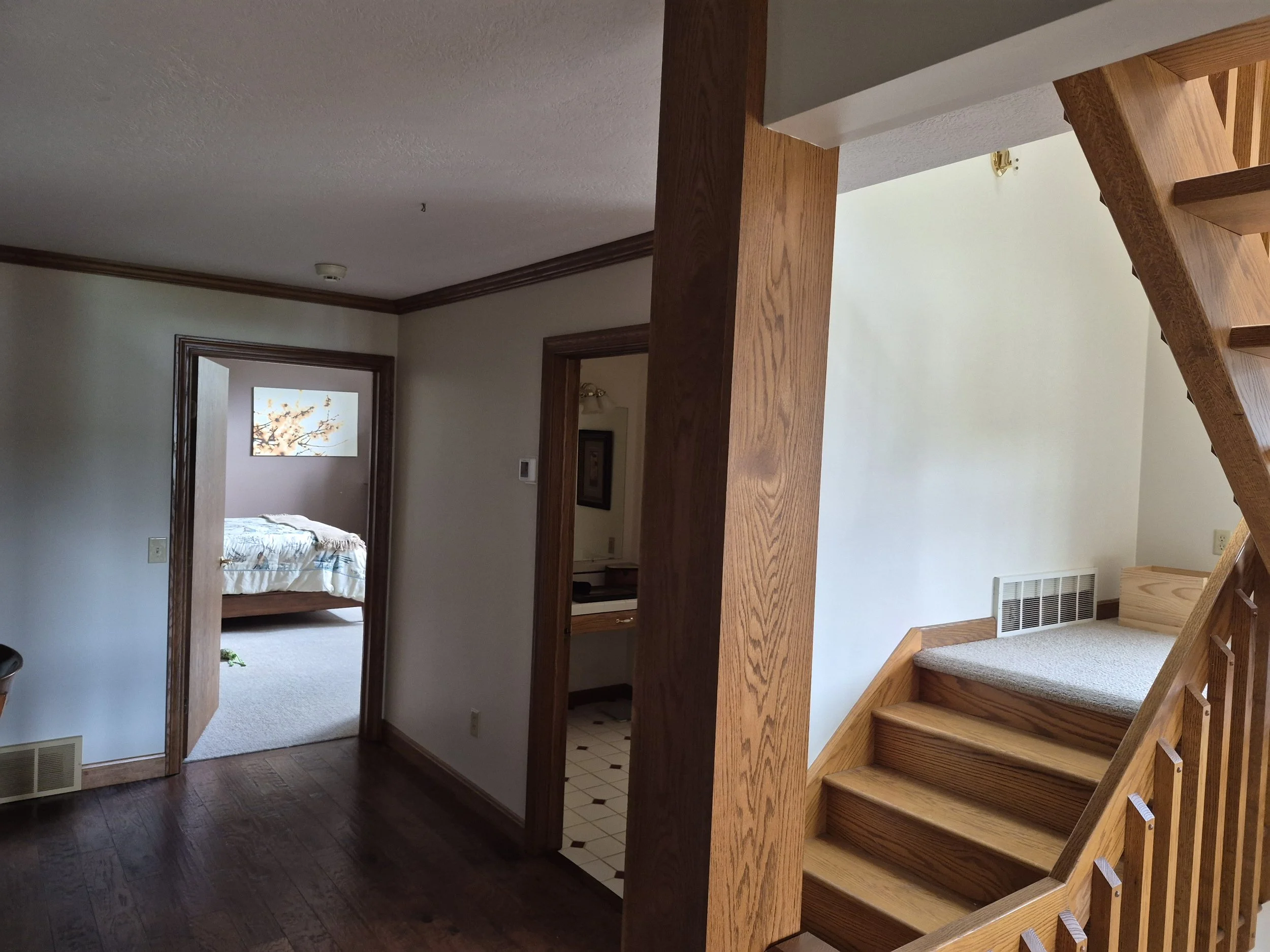 Interior view of a staircase leading to a hallway with a view of a bedroom with a bed and artwork of a tree branch.