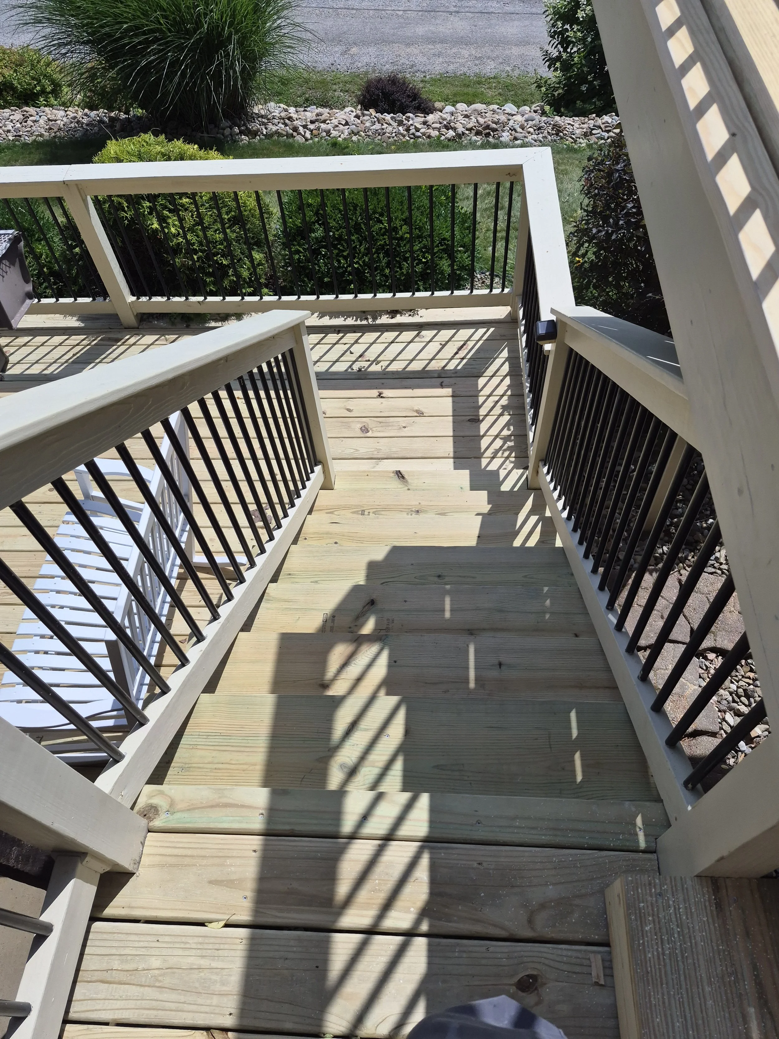 View from the top of a wooden porch or deck, showing the railing, shadows on the wooden floor, and surrounding greenery including bushes, stones, and grass.
