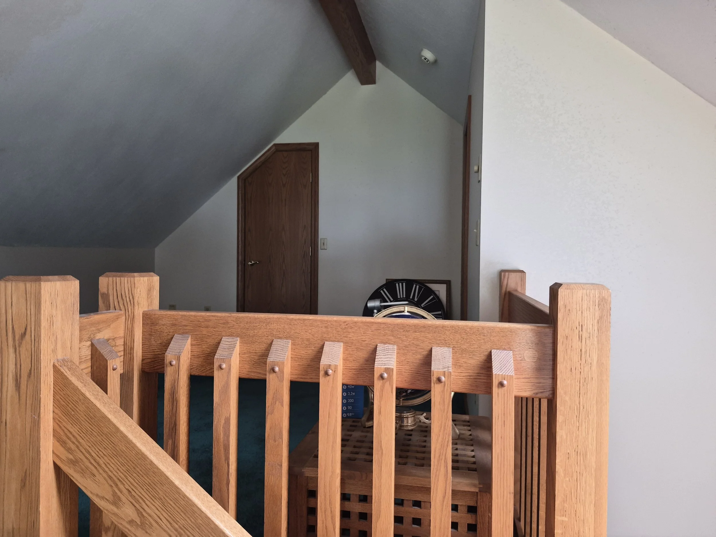 View of a small room or attic space with a sloped ceiling, a wooden door, and a black clock on the wall. There is a wooden railing in the foreground and a storage heater at the bottom of the image.