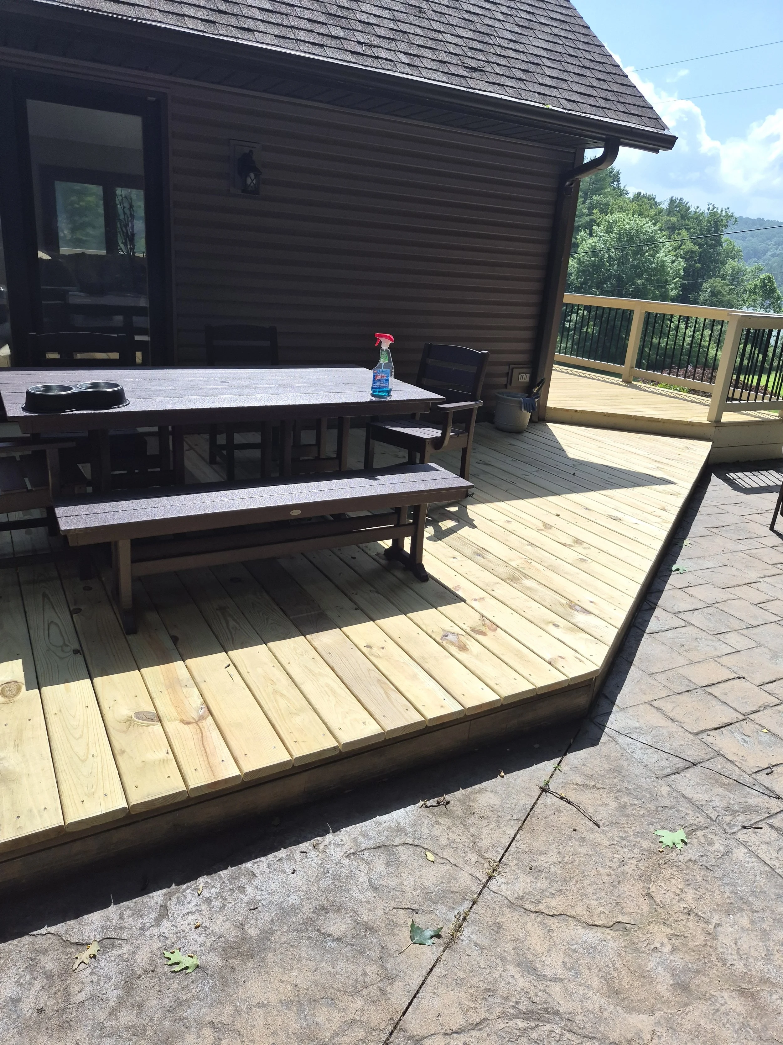 Back deck with new wooden flooring, black outdoor table with two containers, spray bottle, and chairs. House wall with light fixture, sliding glass door, garden and trees in the background.