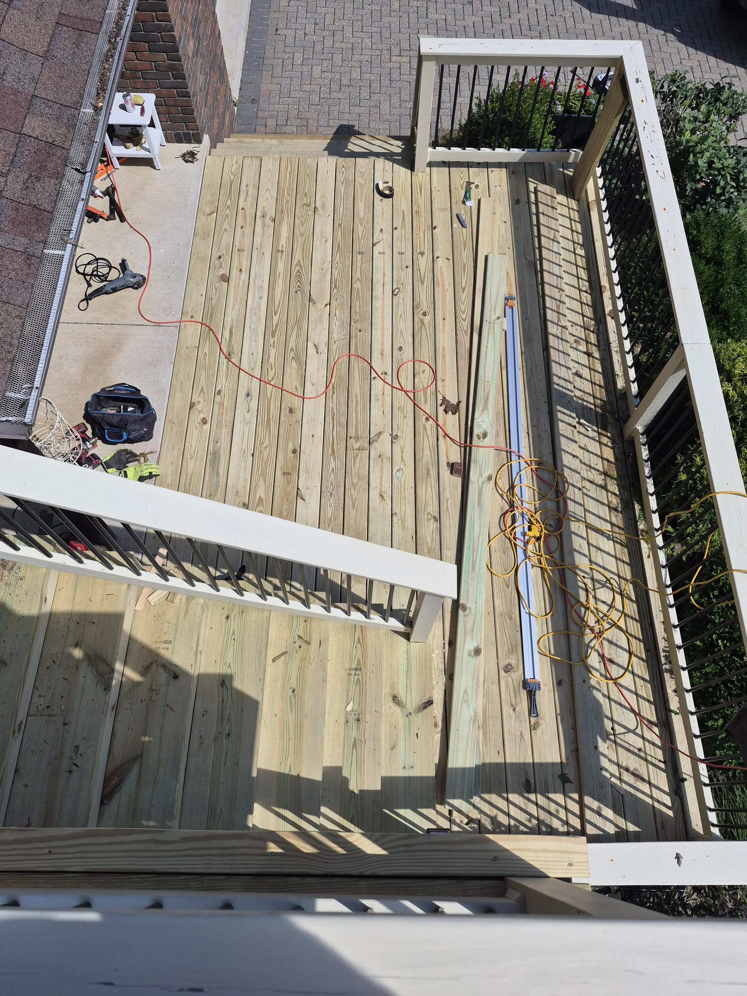A view of a wooden deck in construction with tools, extension cord, and measuring tools placed on it, surrounded by a balcony railing with black metal balusters.
