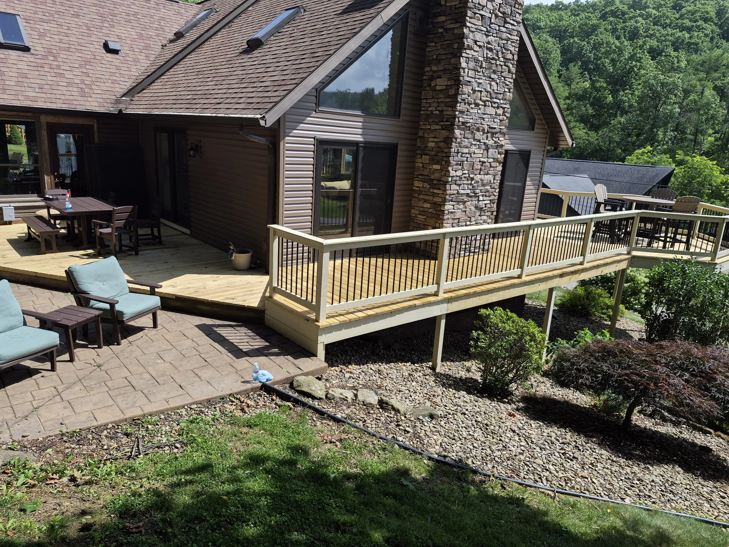 A backyard area with a wooden deck, outdoor dining table with chairs, a stone chimney, and sliding glass doors leading into the house. The yard is surrounded by greenery and trees with some outdoor furniture and a bottle of spray cleaner on the table.