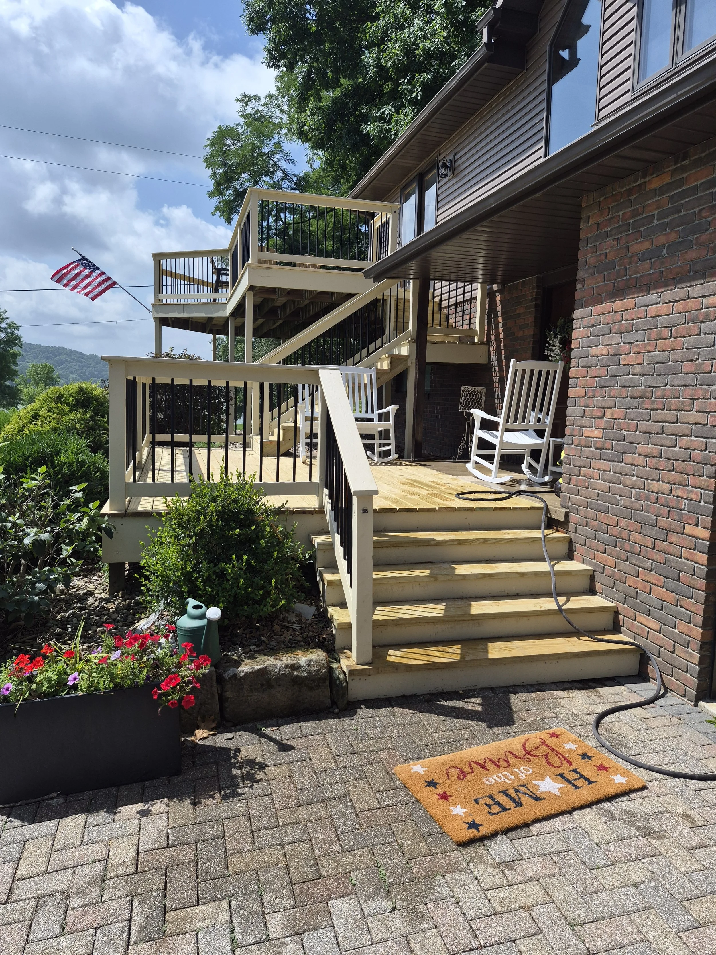 A backyard patio with a wooden deck and staircase, white rocking chairs, potted flowers, and an American flag on a wire. A brick house with windows and a garden with green plants are visible. A colorful Welcome mat is on the brick walkway.
