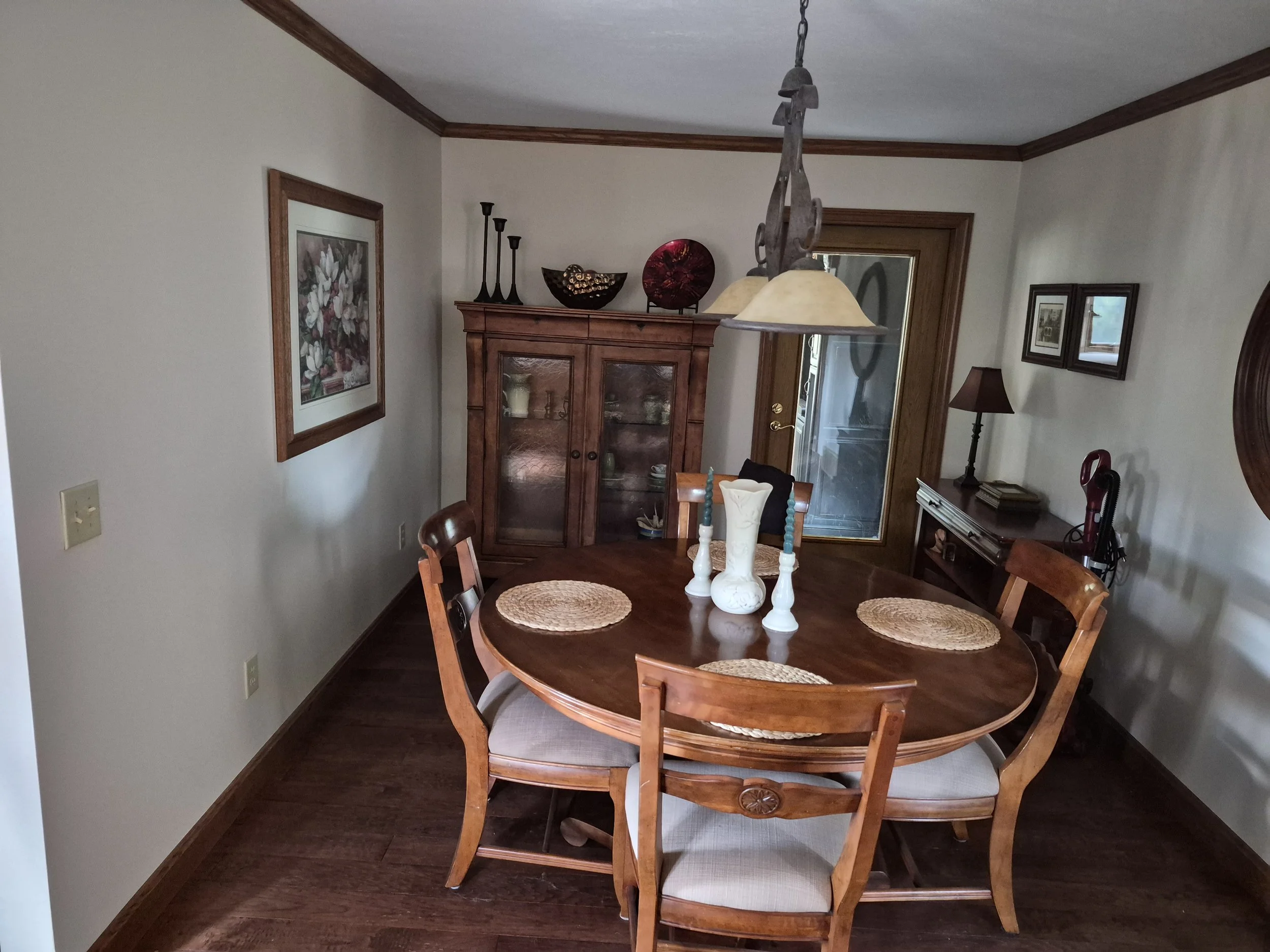 A wooden dining table with six chairs, a decorative white vase with candles, a hanging light fixture, a wooden cabinet with decorative items on top, framed artwork, a small table with a book and a lamp, and a glass door in a dining room.