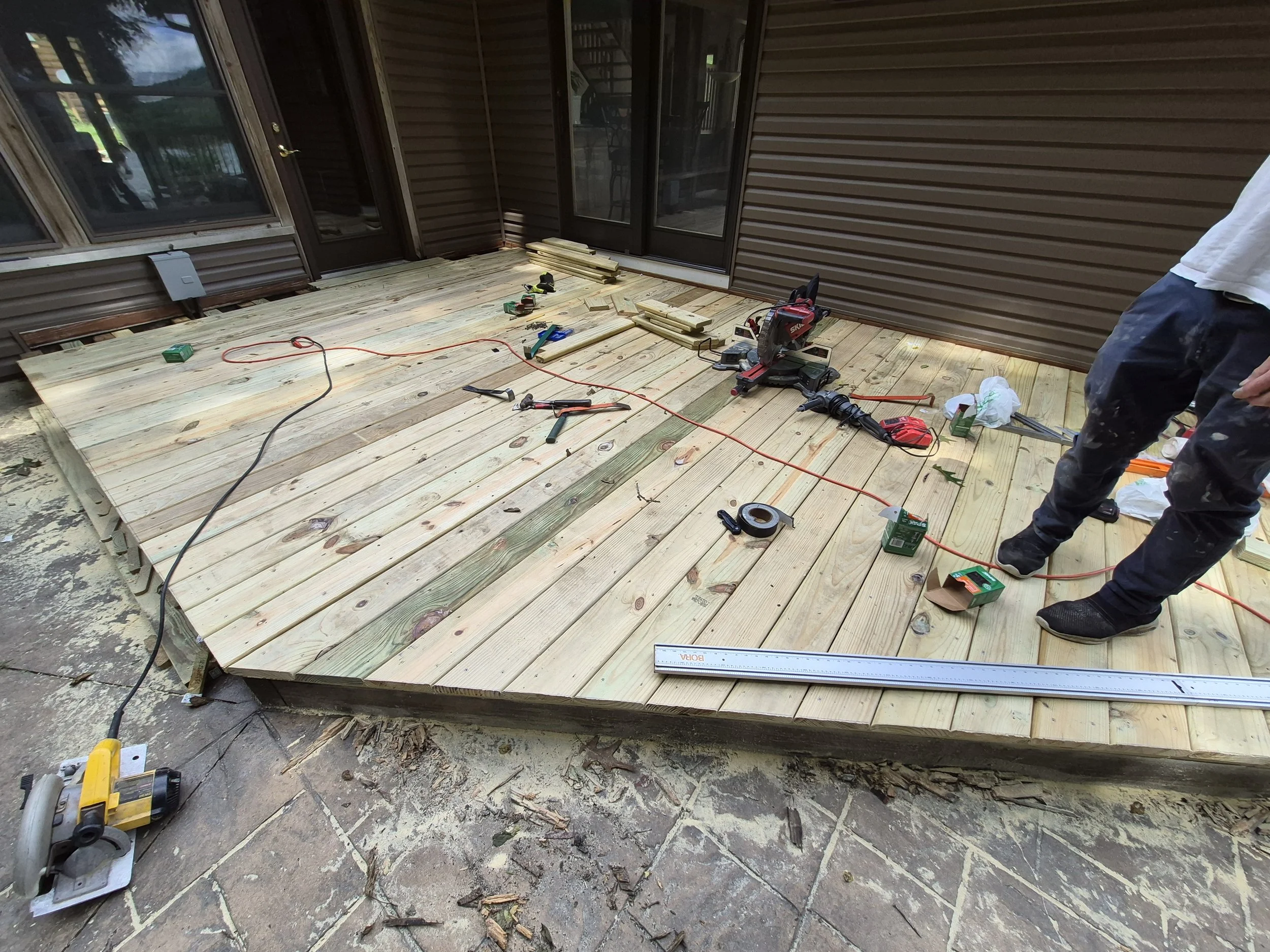 A partially constructed wooden deck with tools and materials on it, attached to a house with brown vinyl siding and large glass doors. A person is standing to the right, wearing dark pants and a white shirt.
