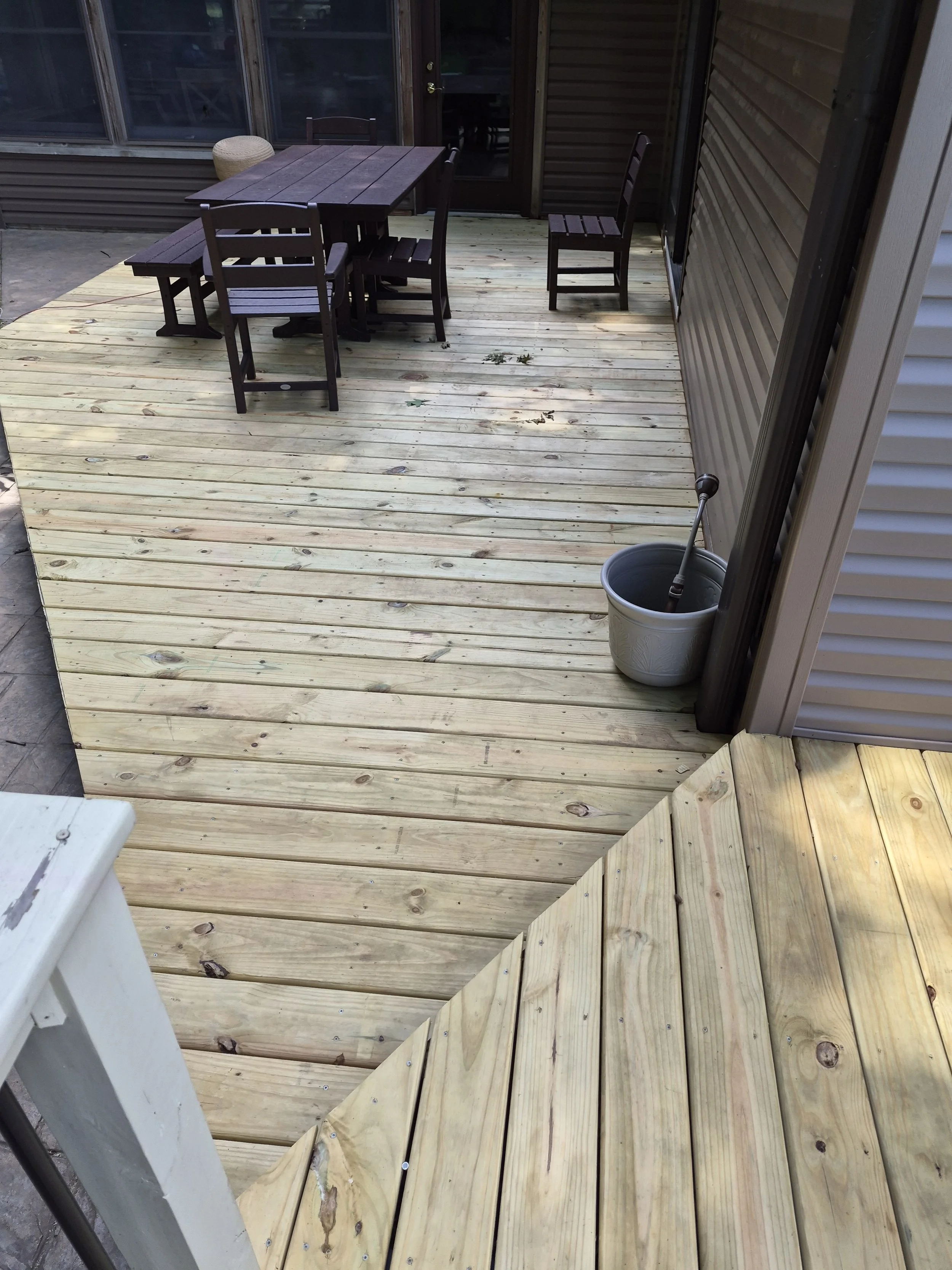 A partially constructed wooden deck with unfinished light-colored wood planks, surrounded by a house with siding, shows a table with chairs, a bucket, a broom, and some leaves and debris on the floor.