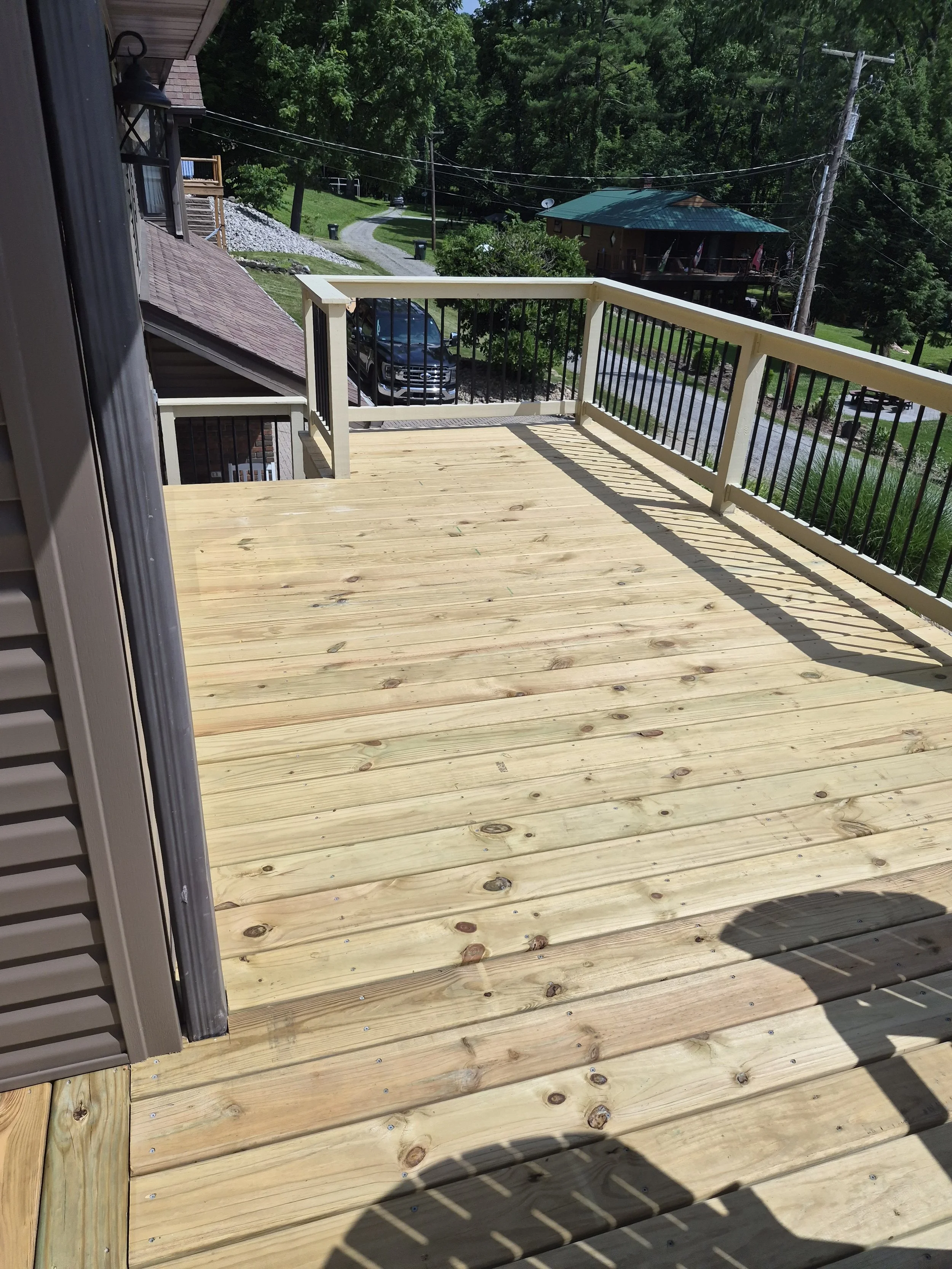 Photo of a wooden balcony with a black railing, overlooking a driveway, a parked truck, a yard with trees, and neighboring houses with trees in the background.